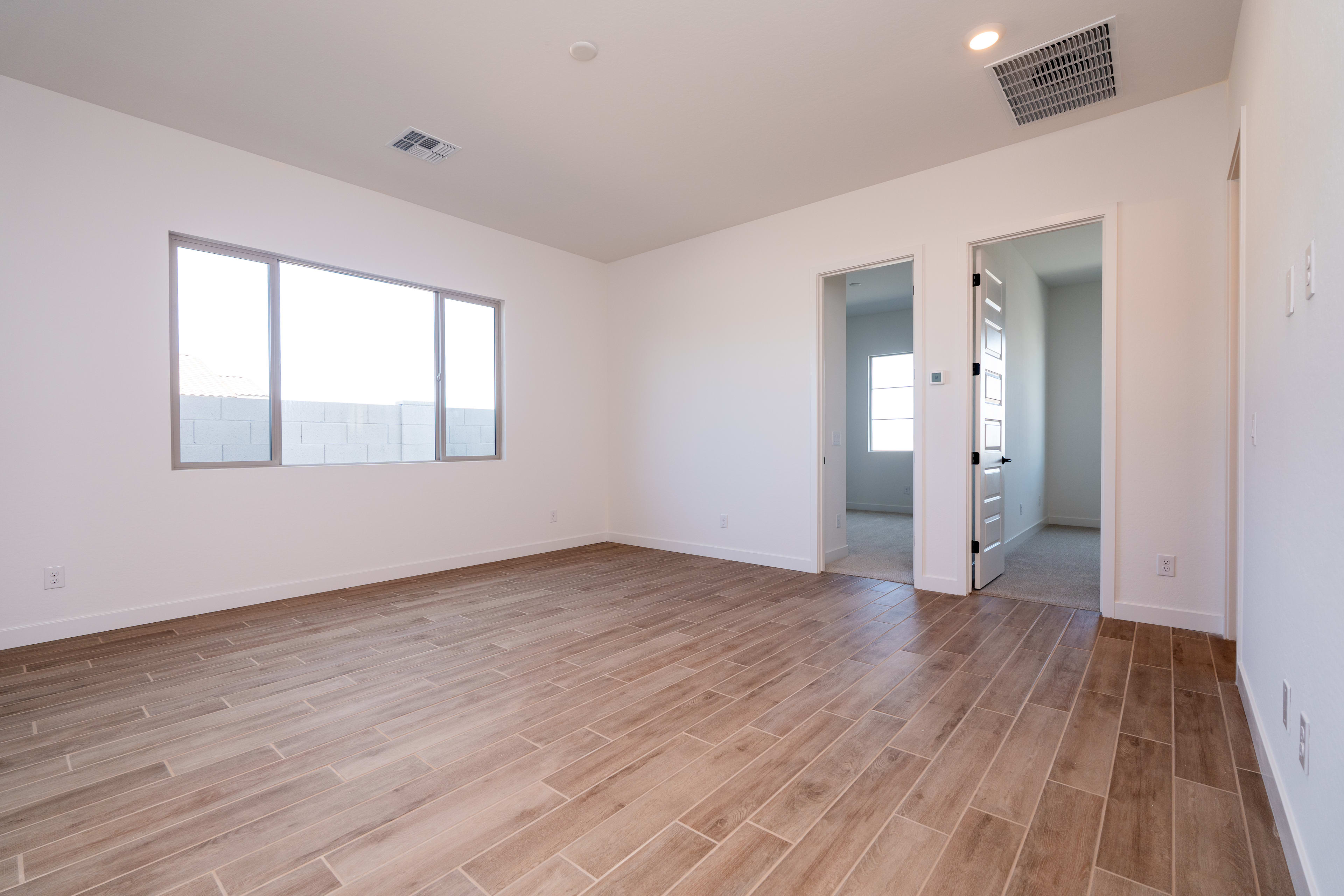 An empty room with white walls, a wooden floor, and a window providing natural light. The room appears to be a bedroom or living space, with a doorway visible in the background.