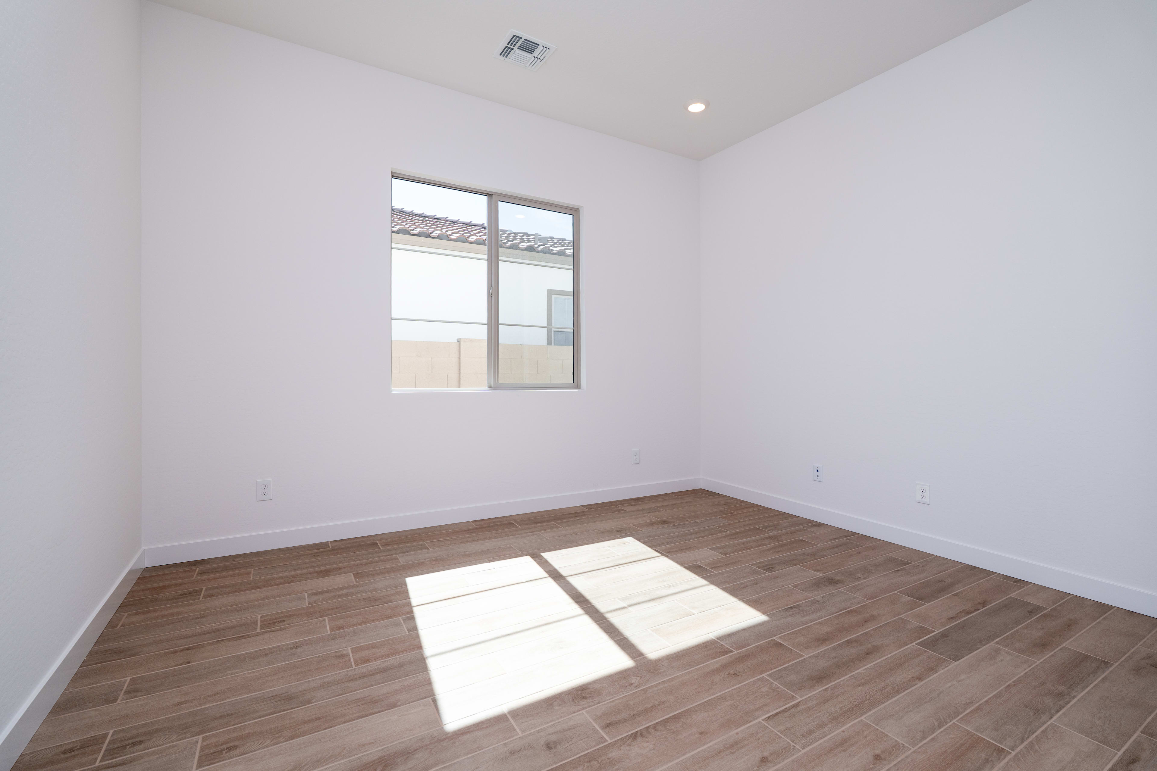 A bright, empty room with white walls and a wooden floor, featuring a window that allows natural light to stream in.
