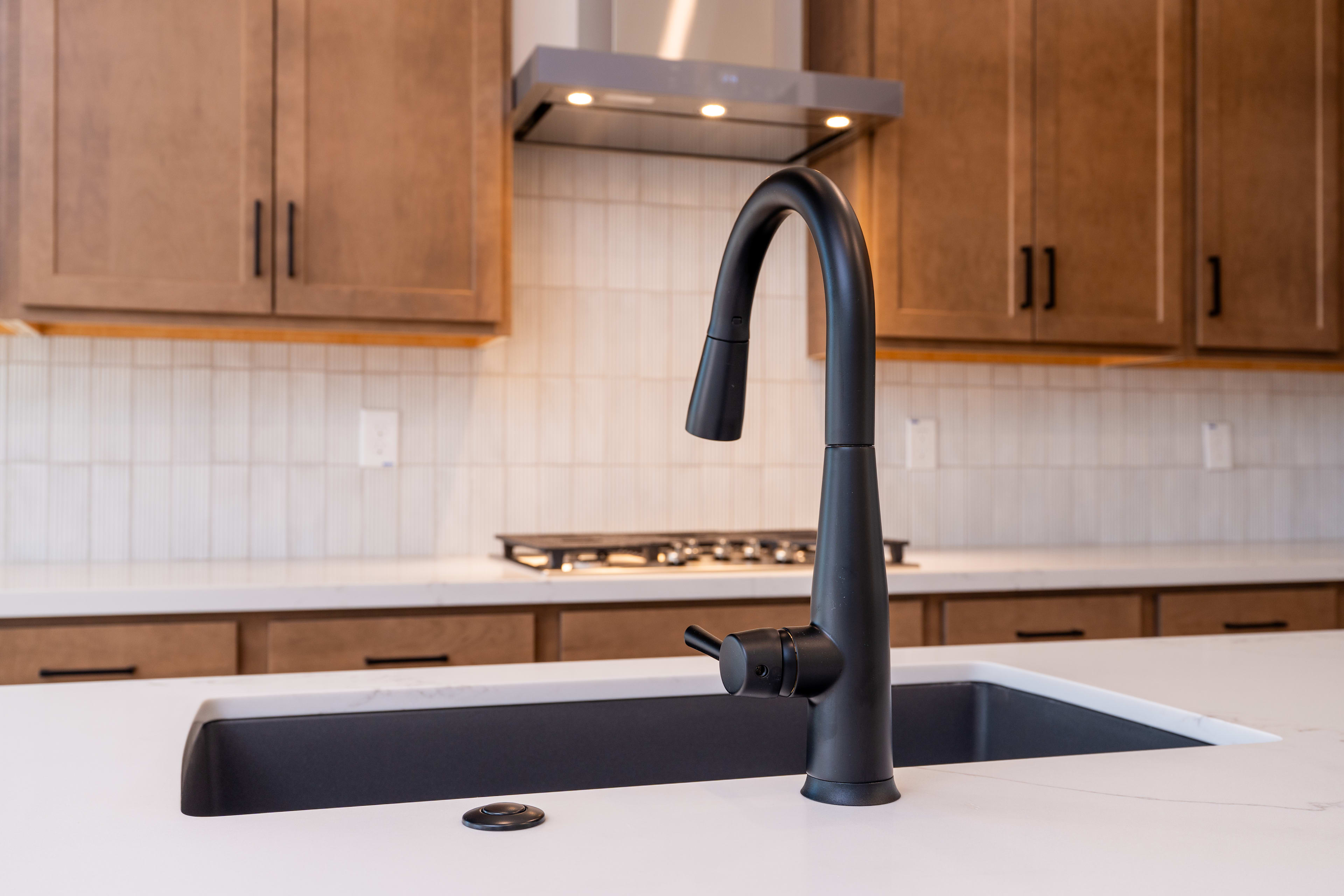 A modern black kitchen faucet with a sleek design sits atop a white sink, surrounded by wooden cabinets in a well-lit kitchen interior.