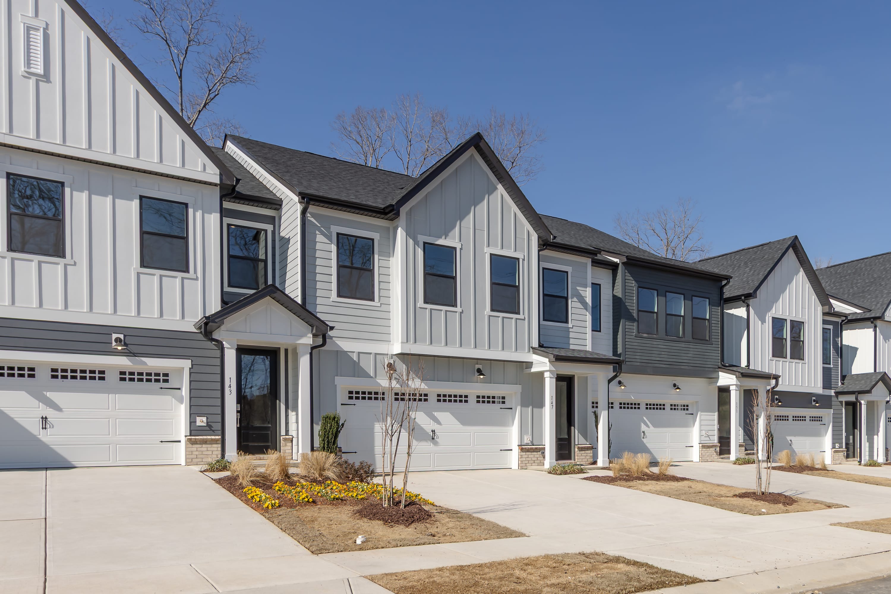 A row of modern, two-story townhouses with white siding, gabled roofs, and attached garages, set against a clear blue sky with bare trees in the background.