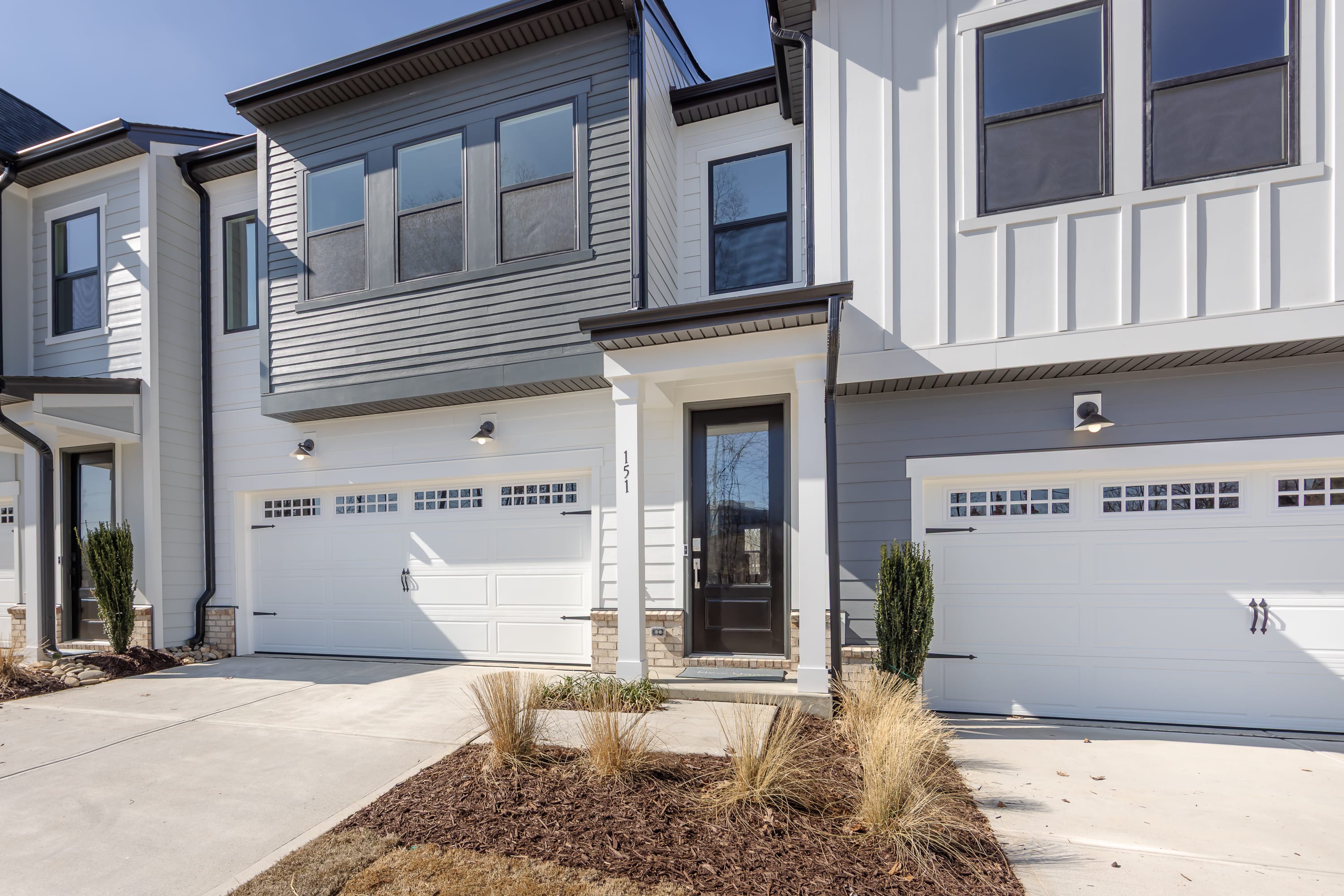 A modern, two-story townhouse with a white exterior, large windows, and a paved driveway leading to a garage door. The front yard features landscaping with grassy areas and decorative plants.