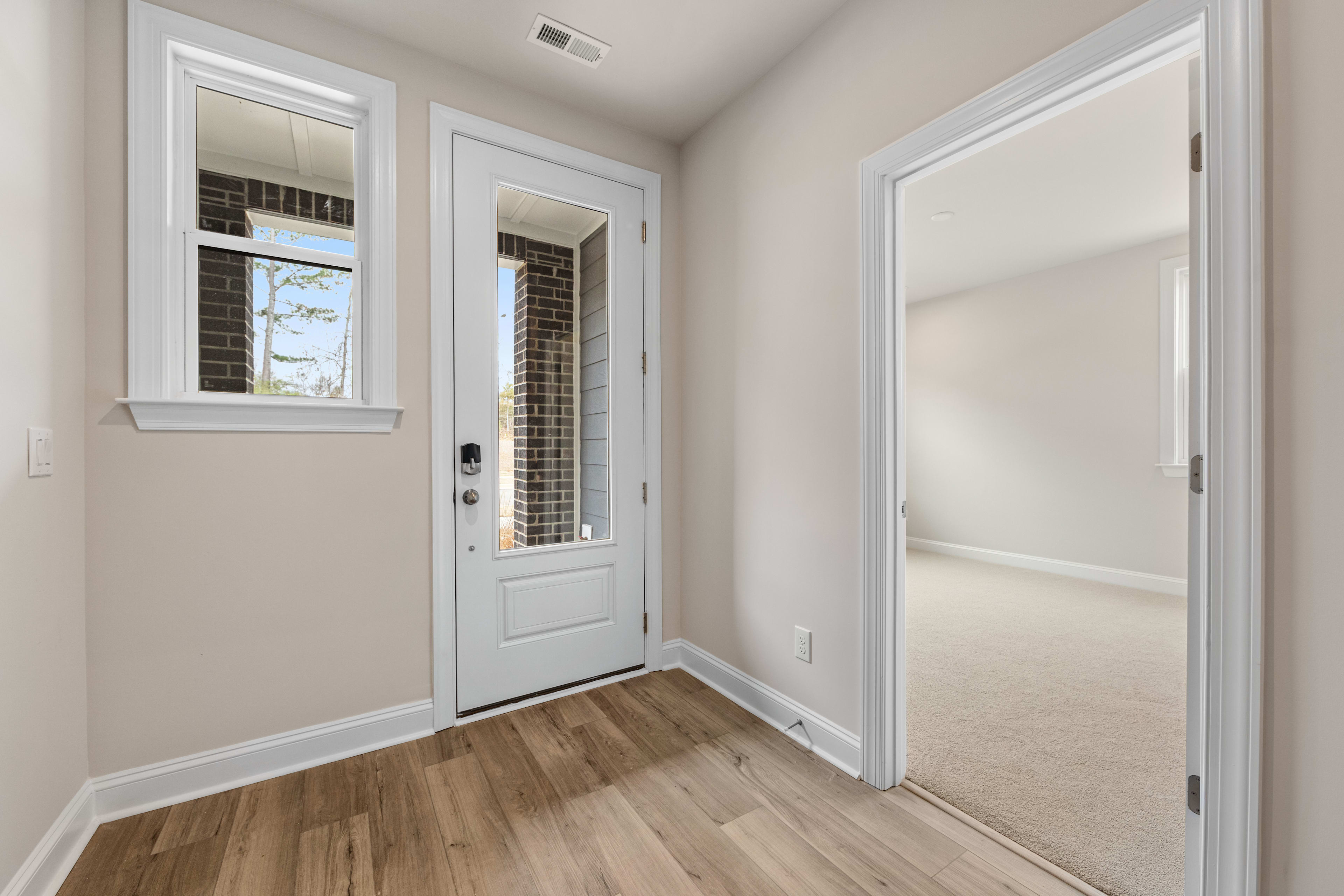 A bright and airy entryway with a white door, windows, and hardwood floors, leading to a hallway and another room.