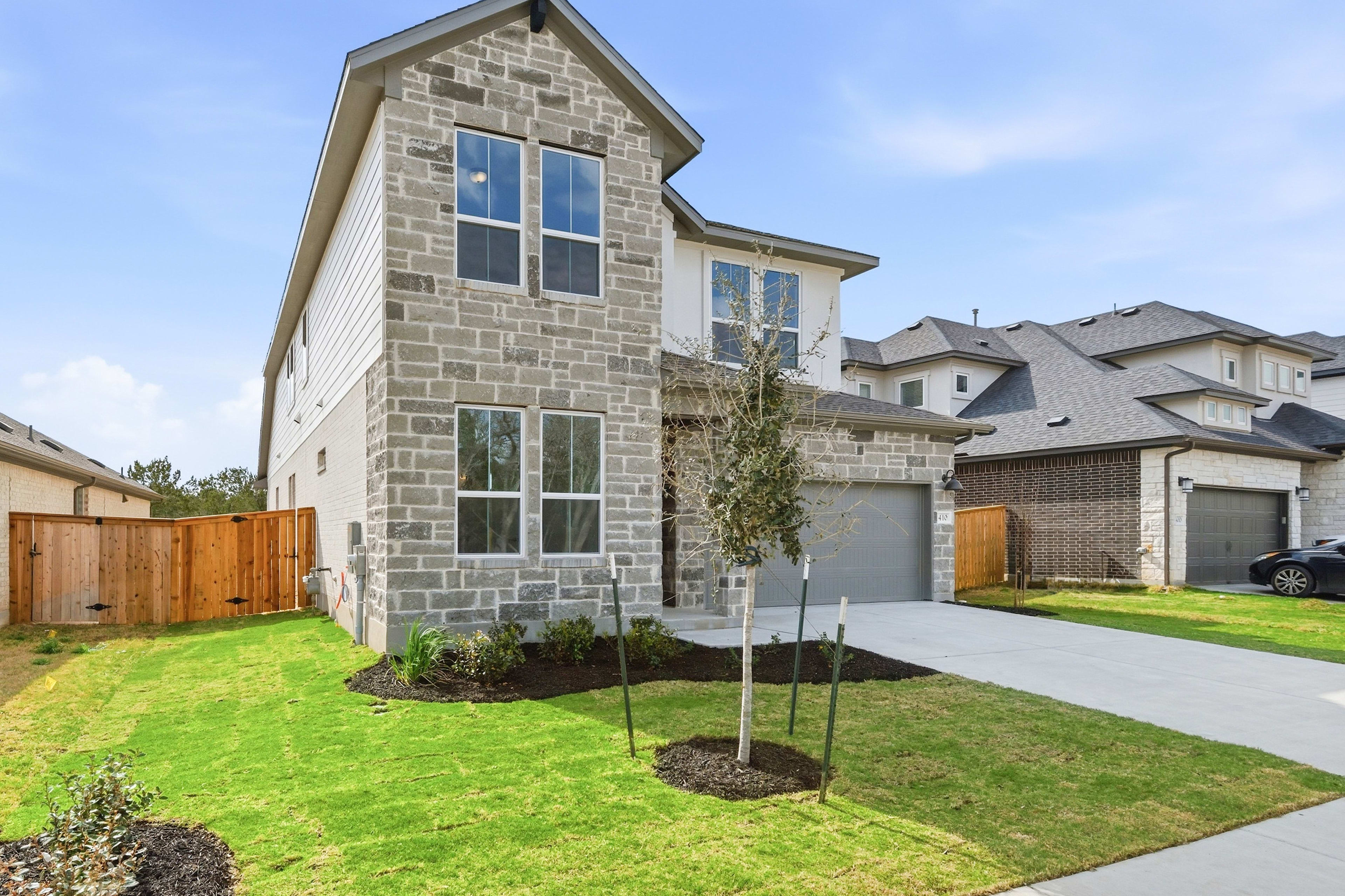 A newly constructed two-story house with a stone exterior, surrounded by a well-manicured lawn and a wooden fence in the foreground, set against a clear blue sky in the background.