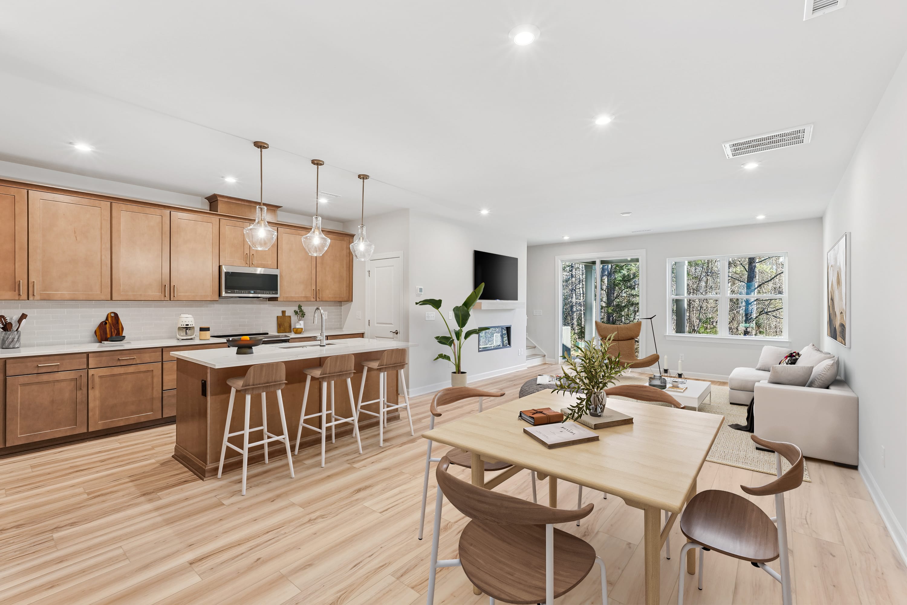 A modern, open-concept kitchen and dining area with wooden cabinetry, pendant lighting, and a cozy seating area with a large window overlooking the outdoors.