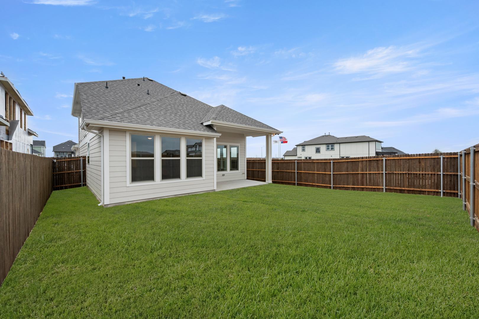 A well-manicured backyard with a lush green lawn, surrounded by a wooden fence, and a small white house with a peaked roof in the background under a clear blue sky.