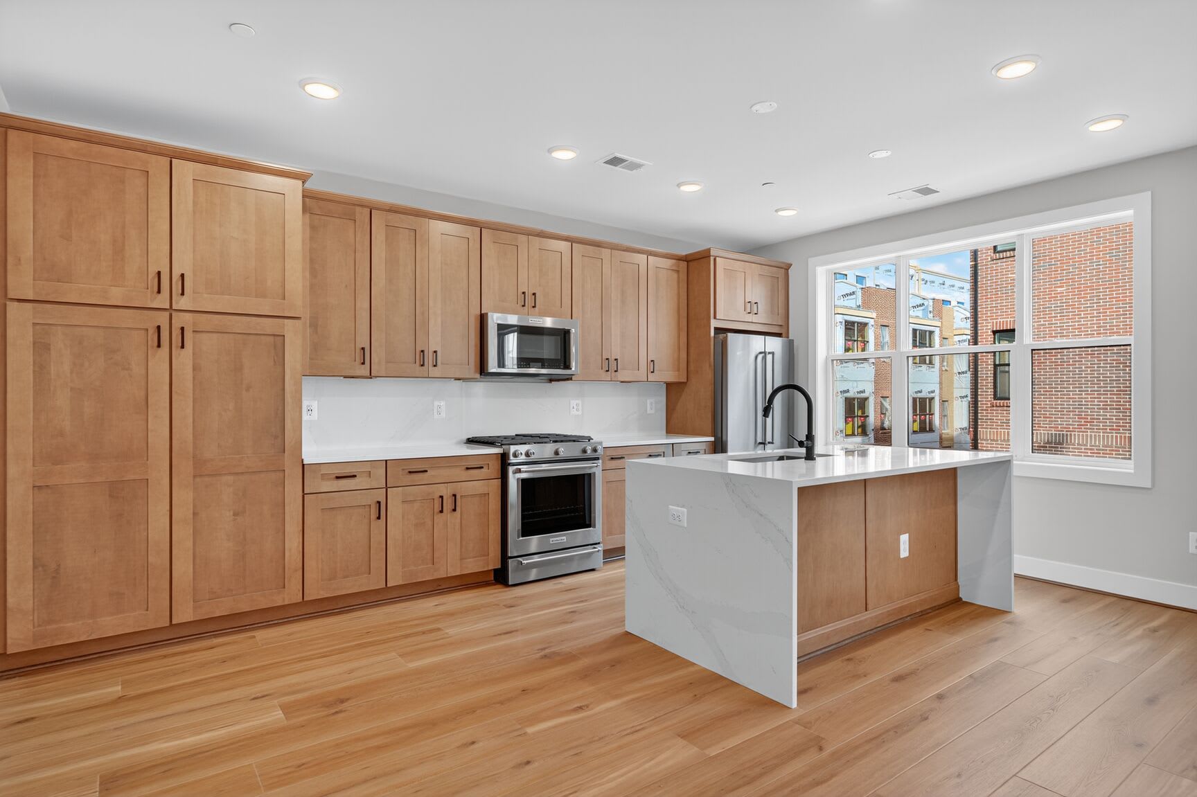 A modern, open-concept kitchen with light wood cabinetry, stainless steel appliances, and a central island with a white countertop, set against a backdrop of large windows allowing natural light to flood the space.