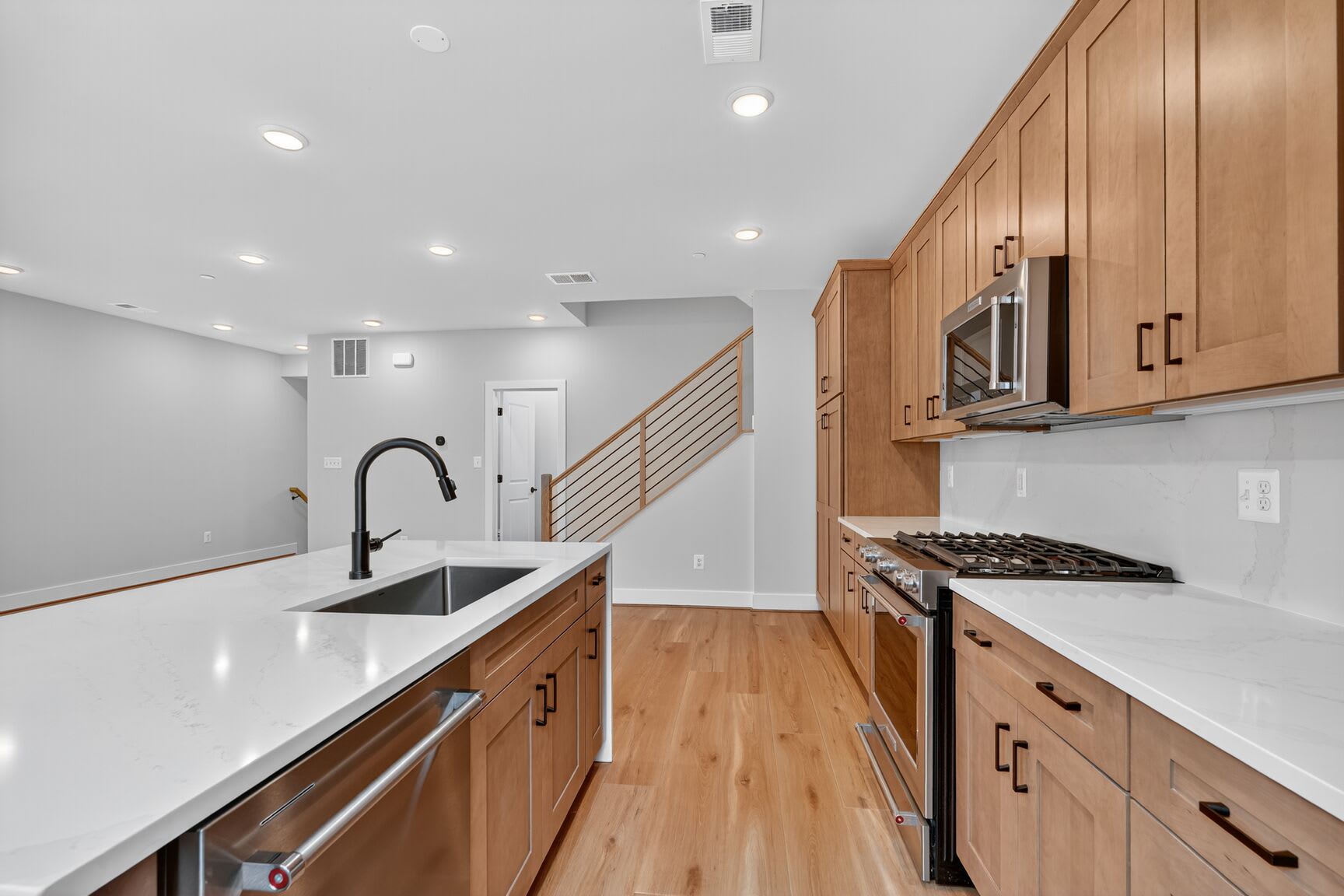 A modern, open-concept kitchen with light wood cabinets, white countertops, and stainless steel appliances, leading into a hallway with a staircase visible in the background.