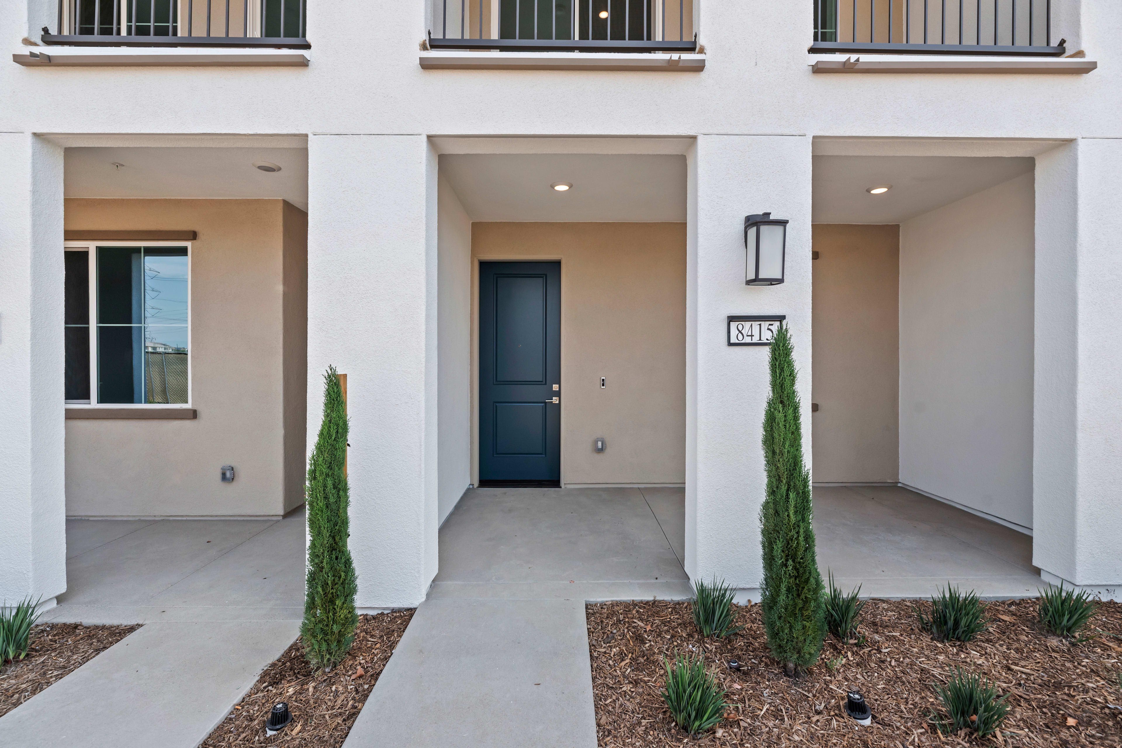 A modern, minimalist townhouse with a clean, neutral exterior, featuring a paved entryway, potted plants, and a dark-colored front door.