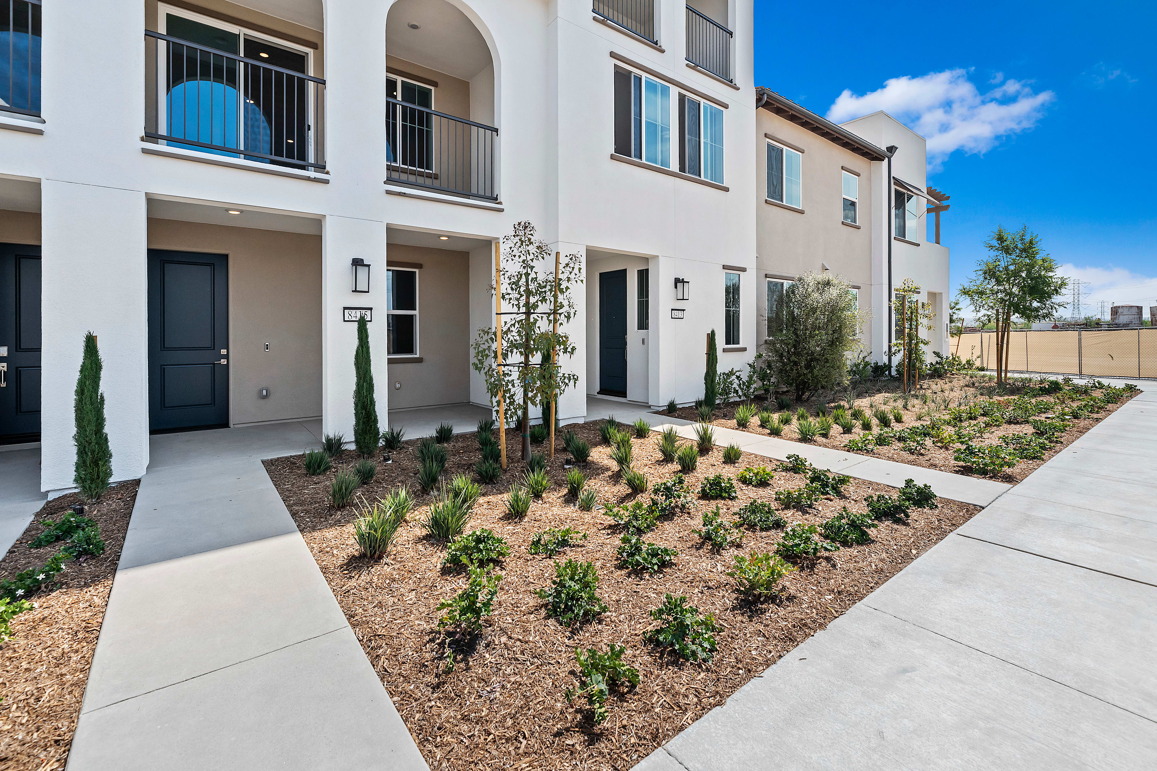 A modern residential building with a well-landscaped front yard featuring a paved walkway, potted plants, and a mix of shrubs and trees.