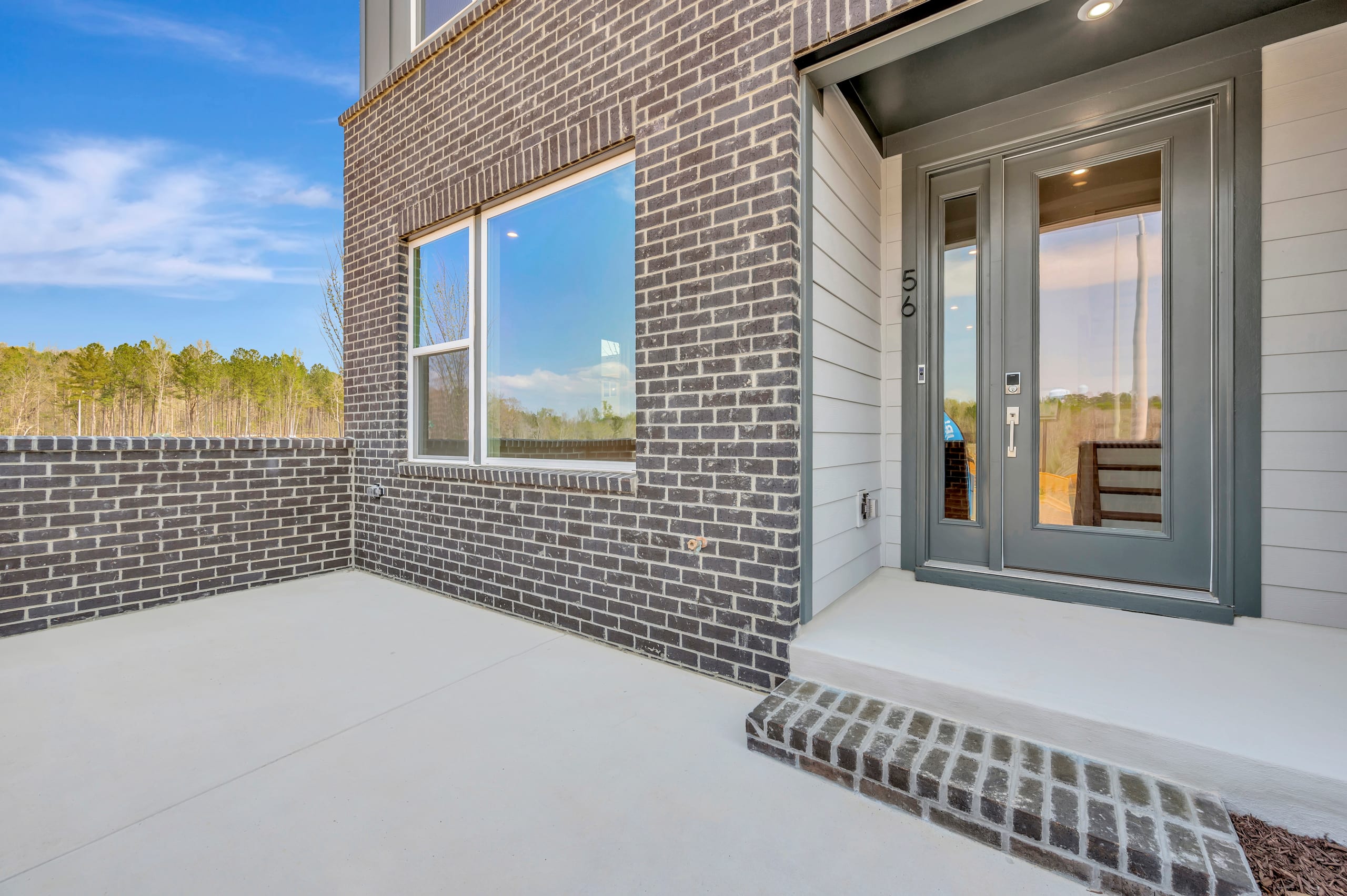 A modern brick and siding exterior of a residential building, with a concrete patio in the foreground and a wooded area visible in the background.