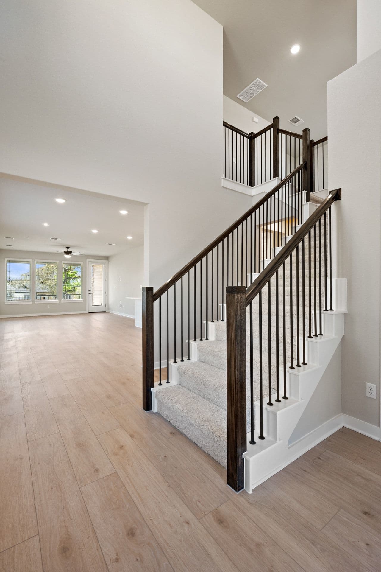 A spacious, open-concept interior with a wooden staircase featuring black metal railings, leading to an upper level. The room has a light, airy feel with hardwood floors and a large window providing natural light.