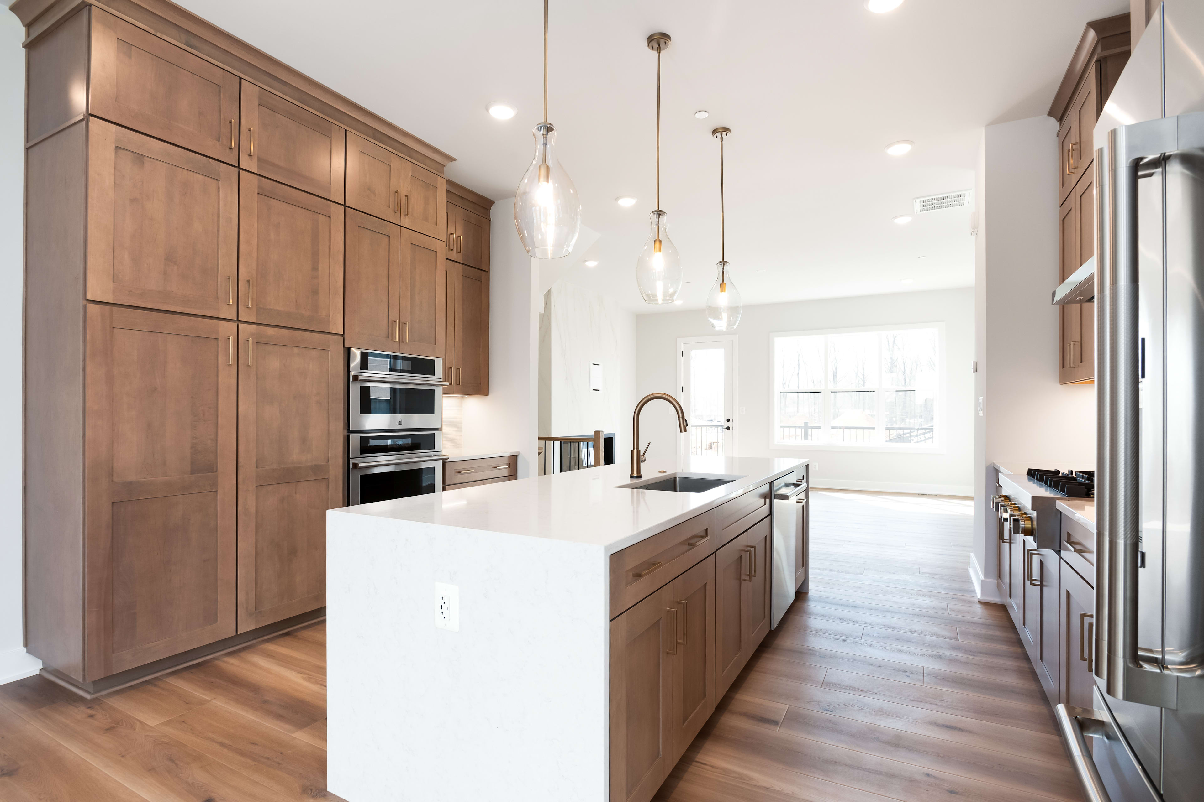 A modern, open-concept kitchen with wooden cabinetry, a white countertop island, and pendant lighting fixtures, set against a bright and airy backdrop.