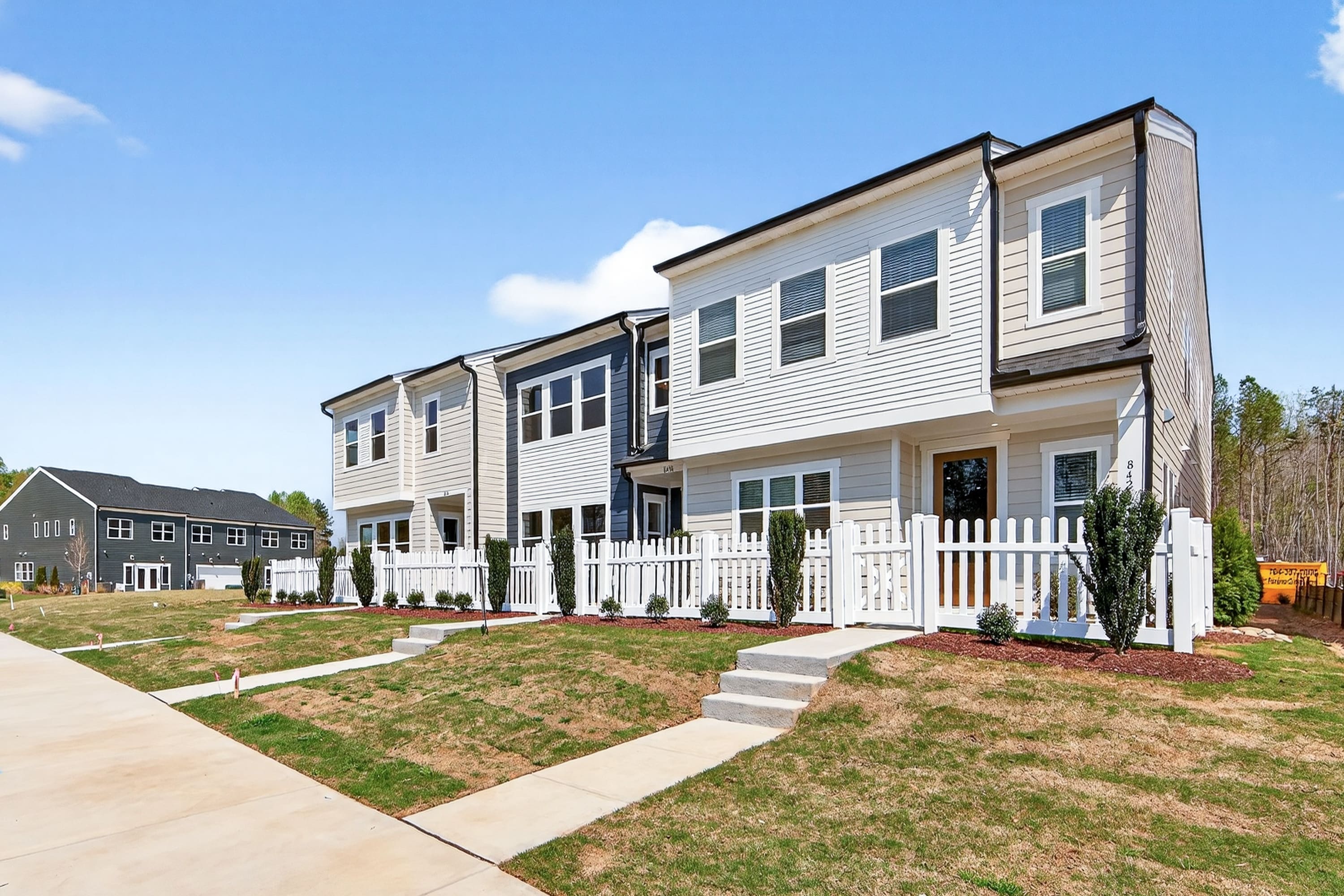 A row of modern, two-story townhouses with white siding and black trim, surrounded by a well-manicured lawn and landscaping, set against a clear blue sky.