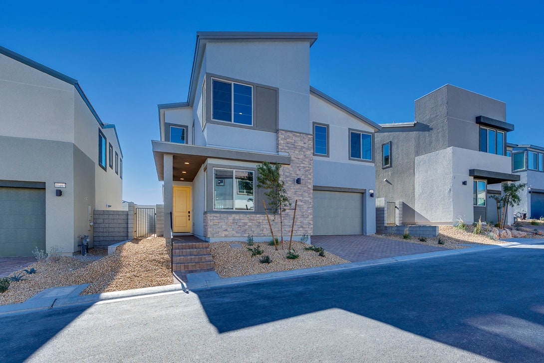 A modern, two-story residential building with a stone facade, large windows, and a paved driveway leading to the front entrance, set against a clear blue sky.