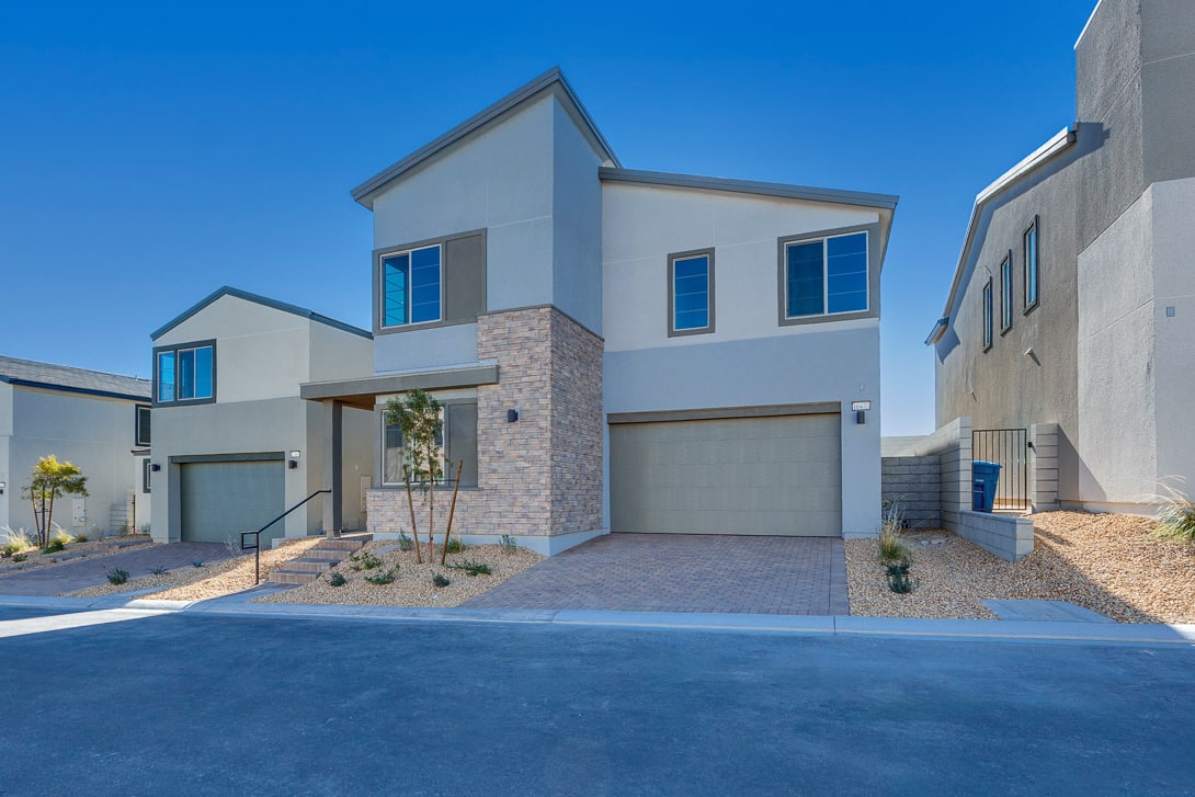 A modern, two-story residential building with a stone facade, a garage, and a paved driveway, set against a clear blue sky.