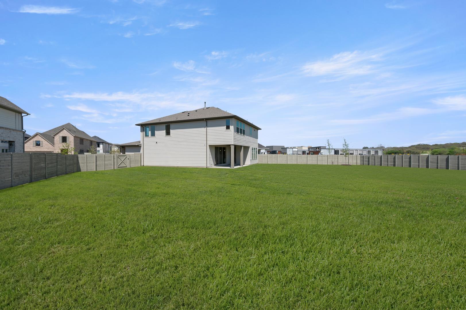 A well-manicured lawn stretches out in the foreground, leading to a two-story white house with a peaked roof in the background, surrounded by a fenced-in yard.
