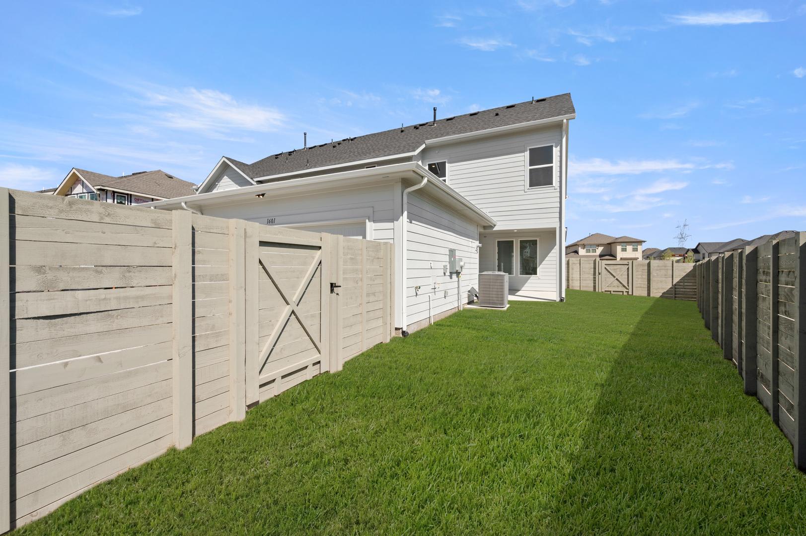 A well-maintained residential neighborhood with a grassy yard, wooden fences, and a two-story house with a white exterior and a slanted roof.