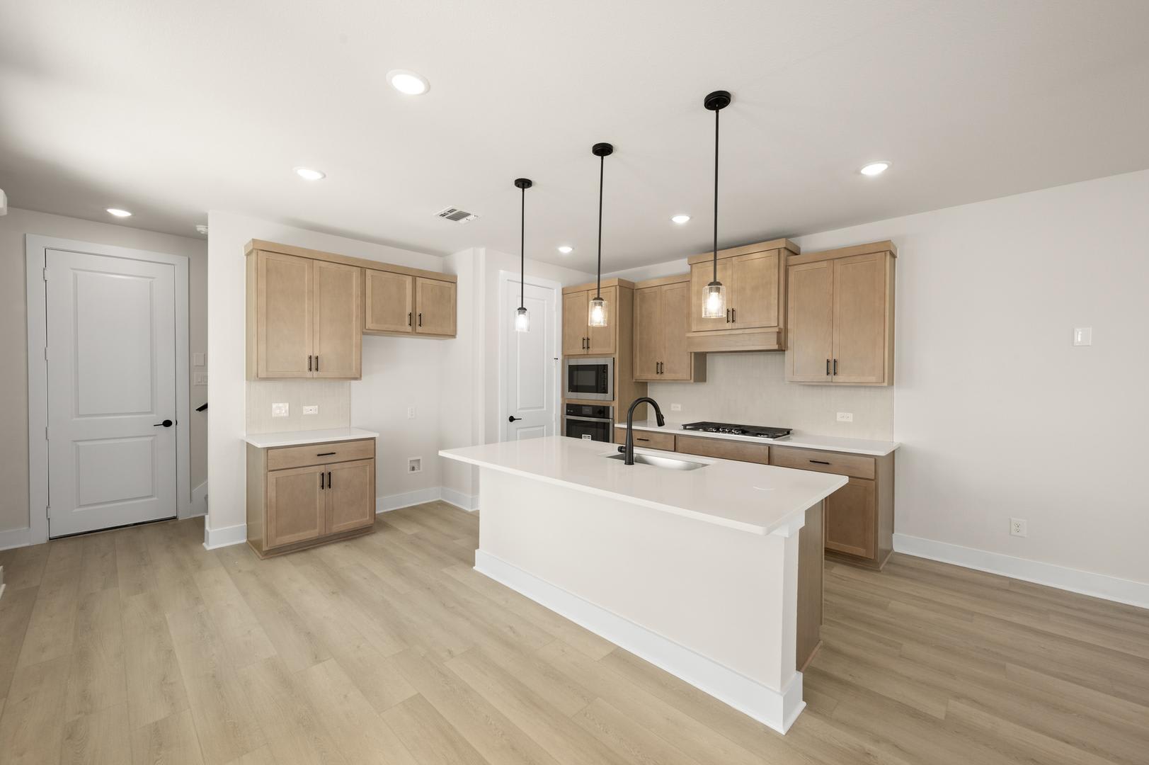 A modern, open-concept kitchen with light-colored wood cabinets, a white countertop, and pendant lighting fixtures hanging from the ceiling.