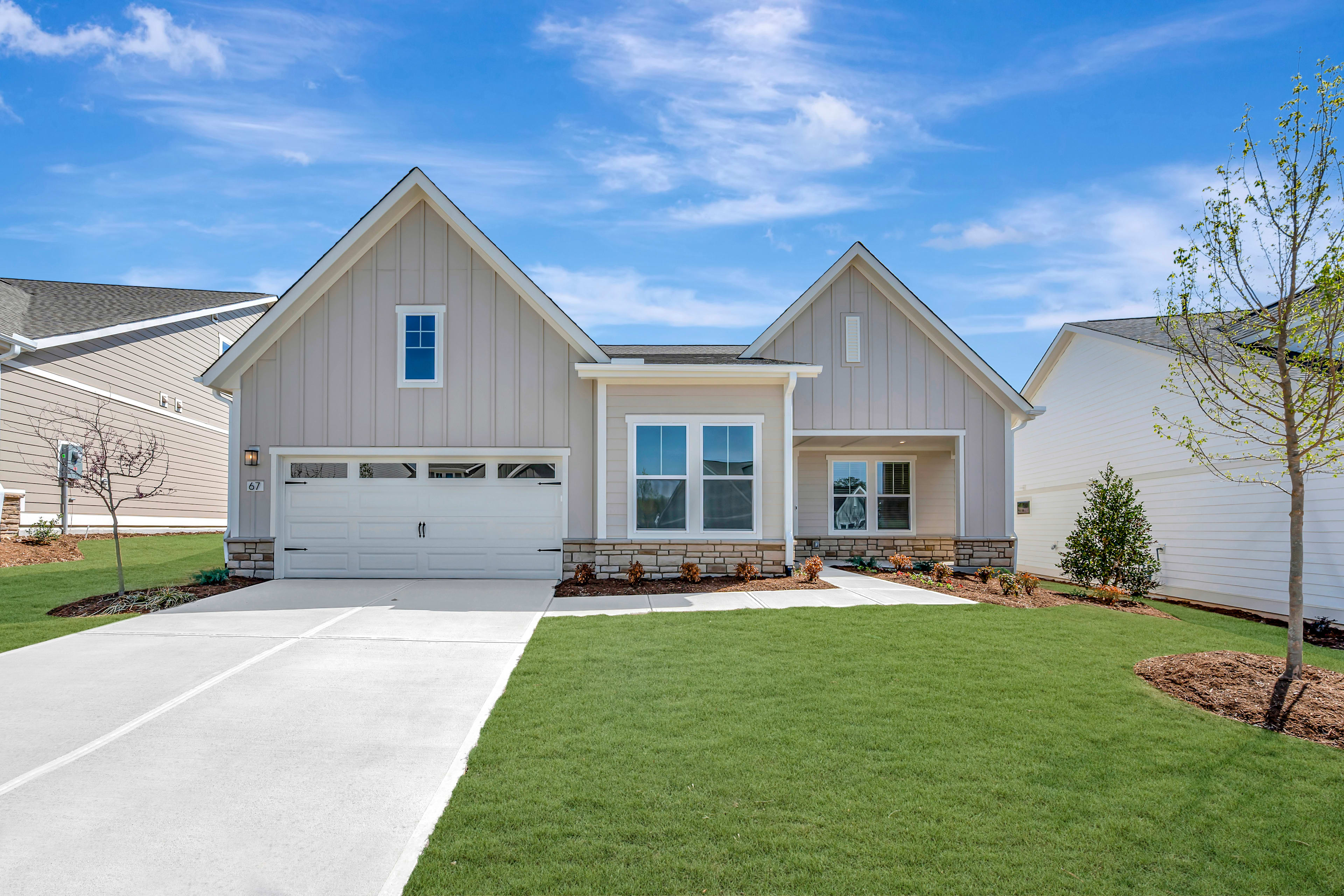A well-manicured lawn surrounds a modern, two-story house with a gabled roof, white siding, and a garage door, set against a clear blue sky with scattered clouds.