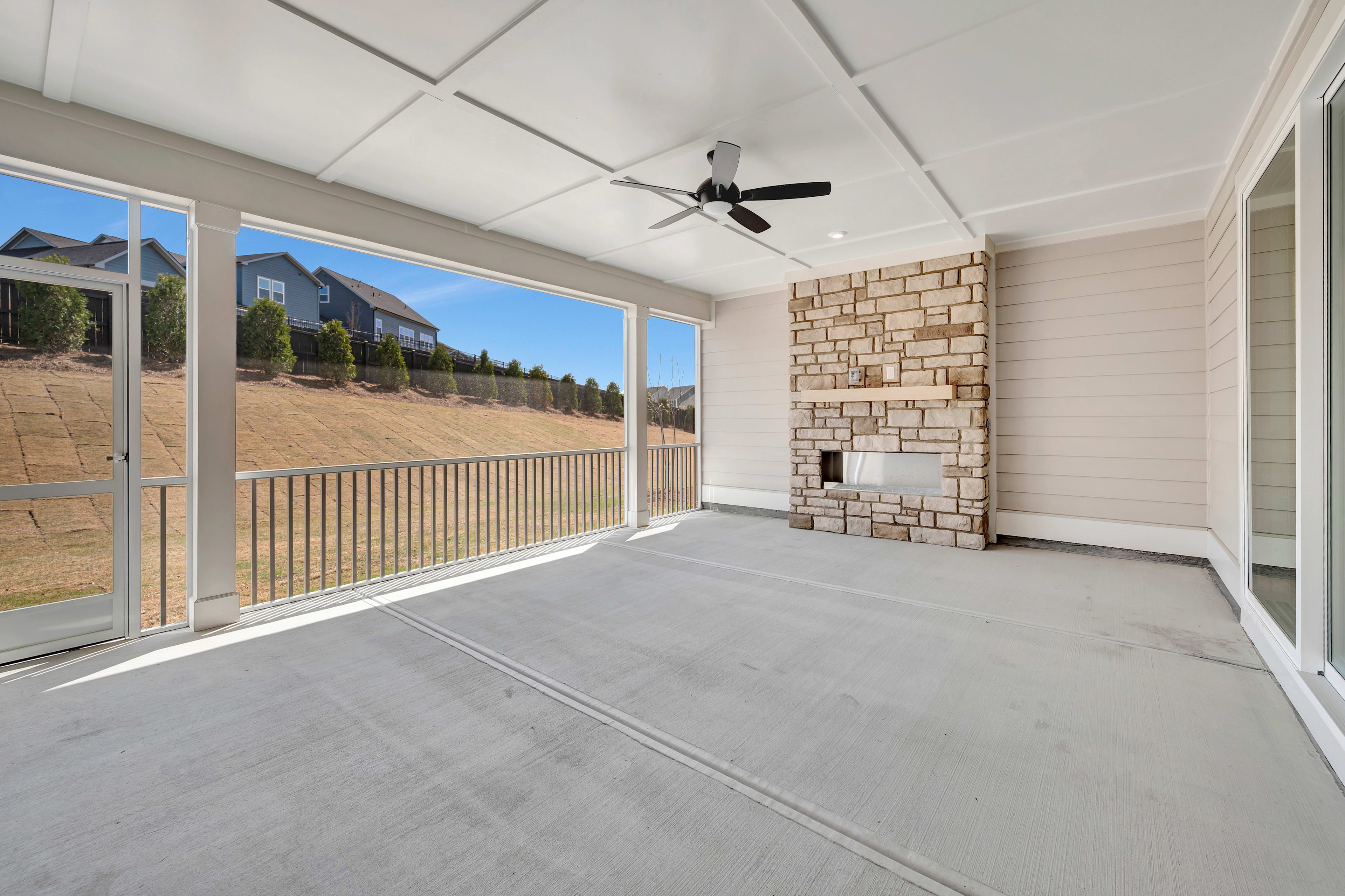 A spacious covered patio with a ceiling fan, surrounded by large windows overlooking a residential neighborhood with houses and trees in the background.