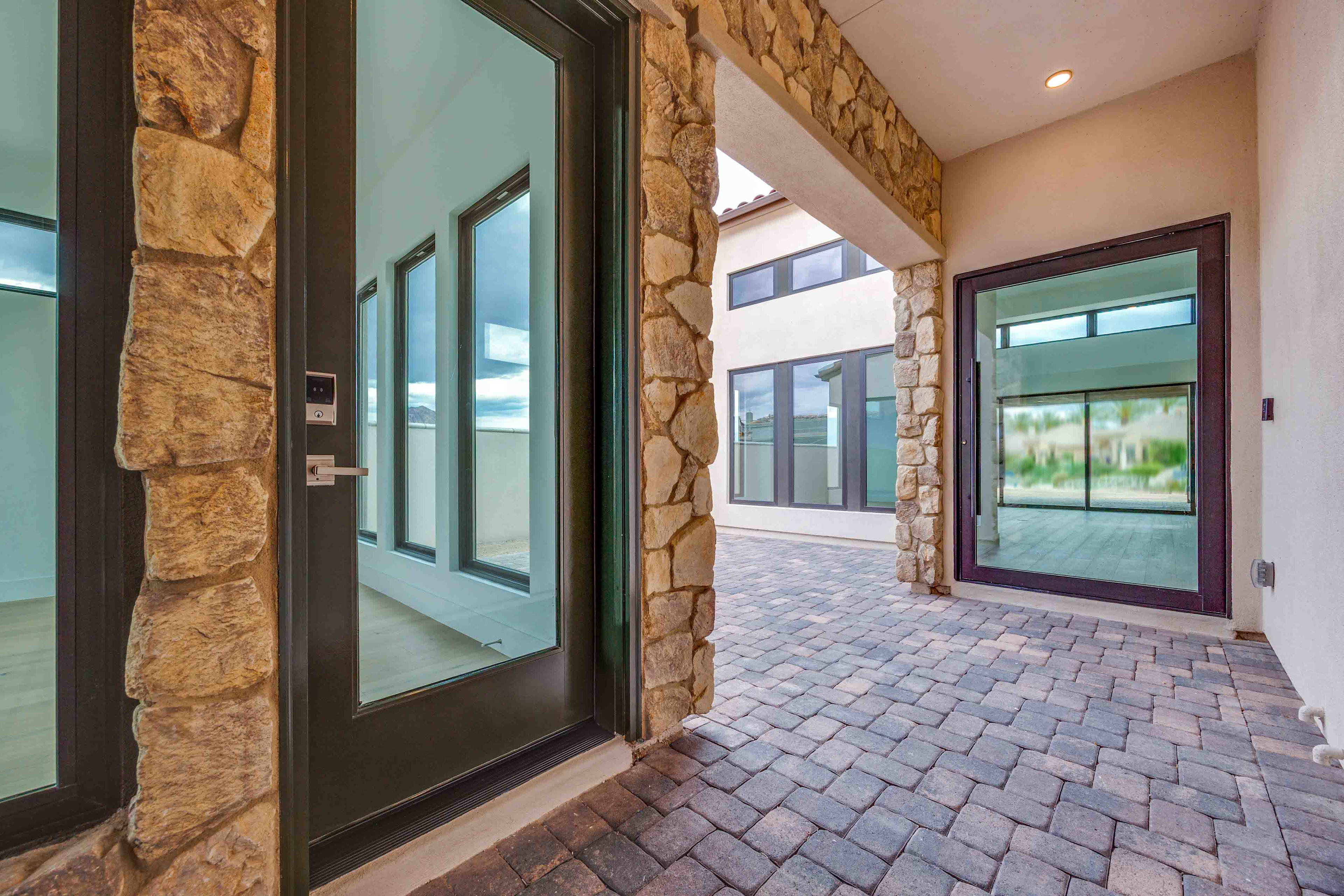 The image depicts a spacious entryway with a stone-clad wall, large glass windows, and a brick-paved floor leading to an interior space.