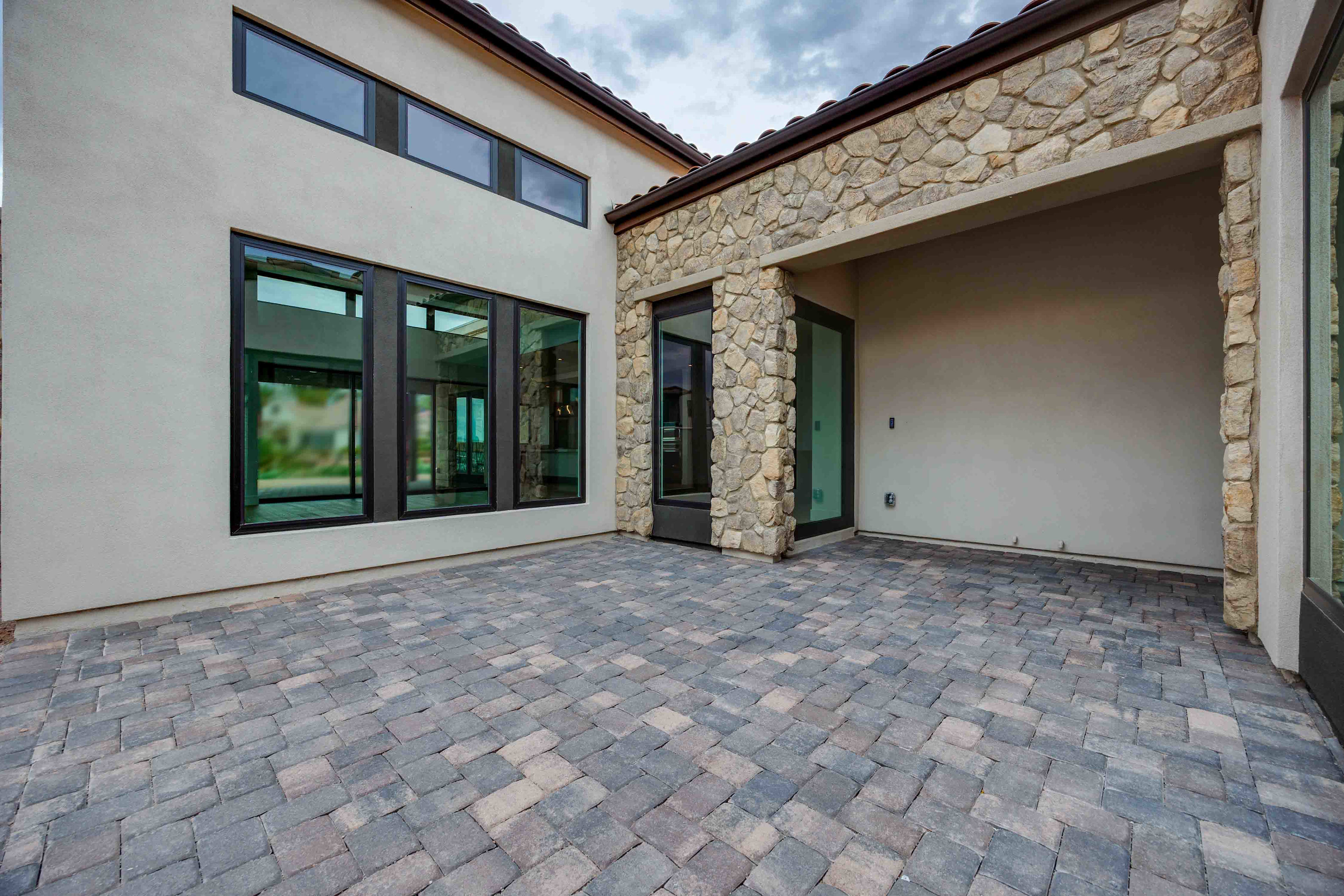 The image depicts a modern, stone-clad exterior of a residential building, with a paved courtyard in the foreground and large windows providing ample natural light.