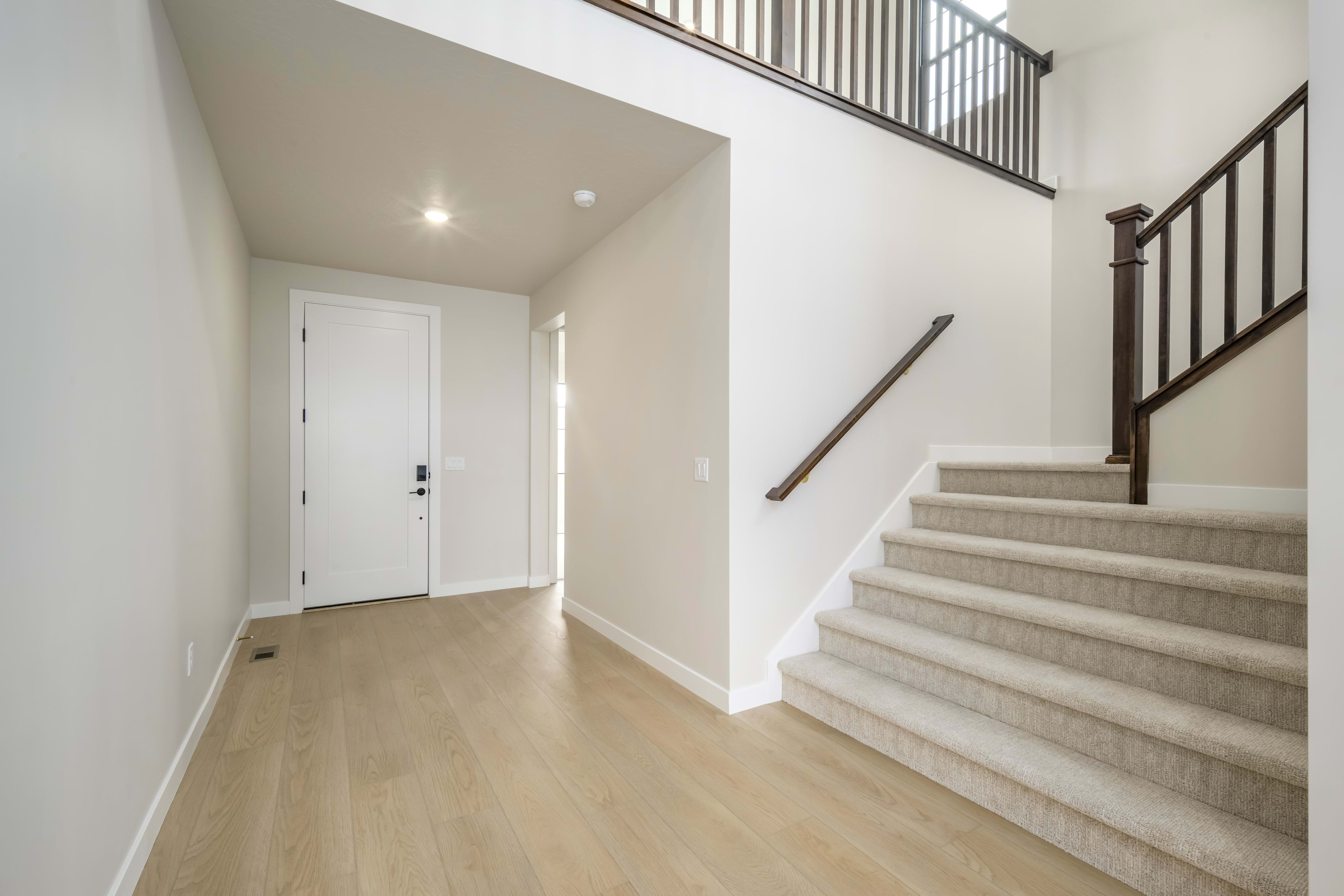 A bright and spacious hallway with a staircase leading up, featuring a white door and hardwood floors.