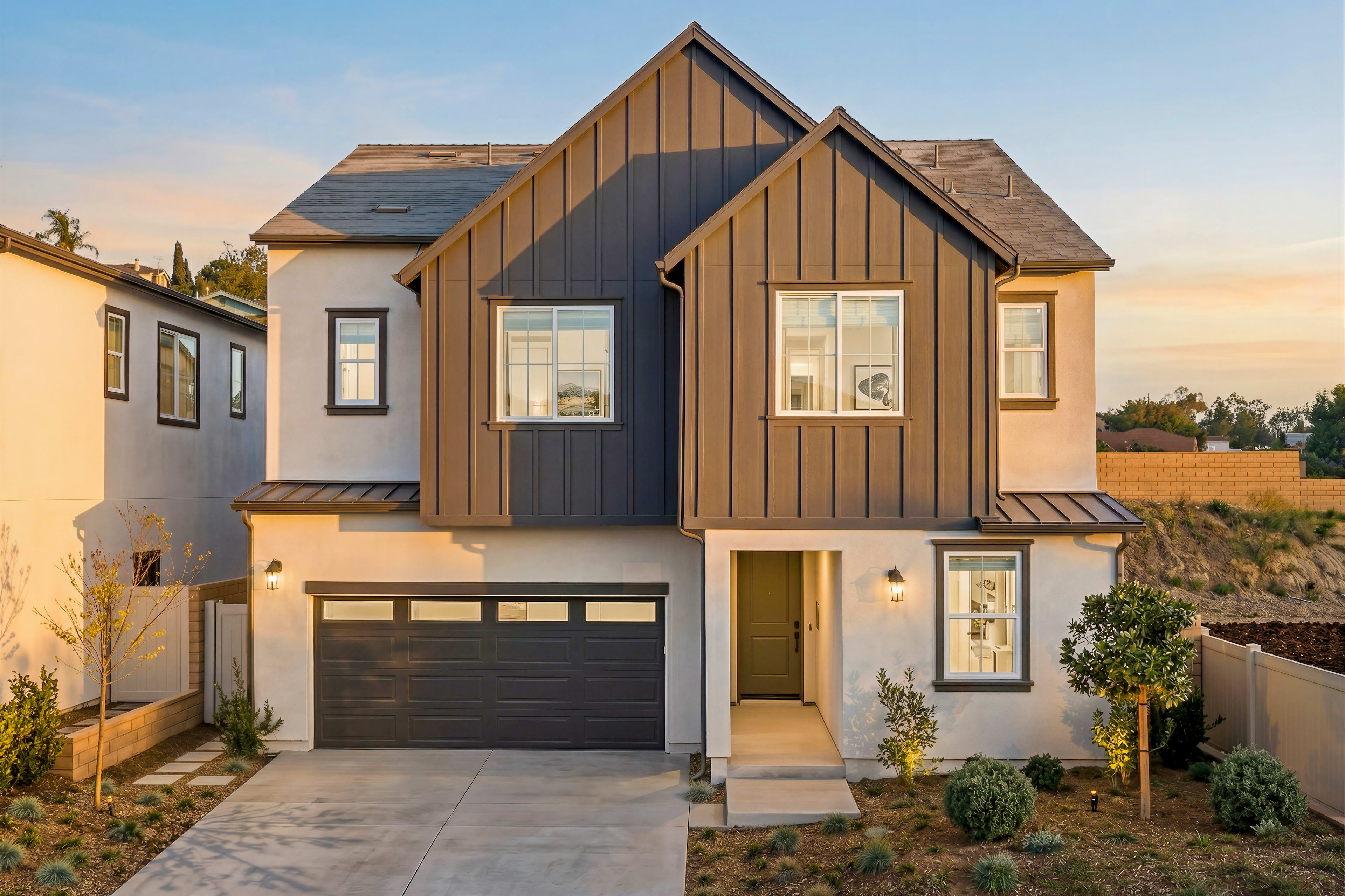 A modern two-story house with a mix of wood and gray siding, a garage door, and a paved driveway in the foreground, set against a backdrop of a clear sky and lush greenery.