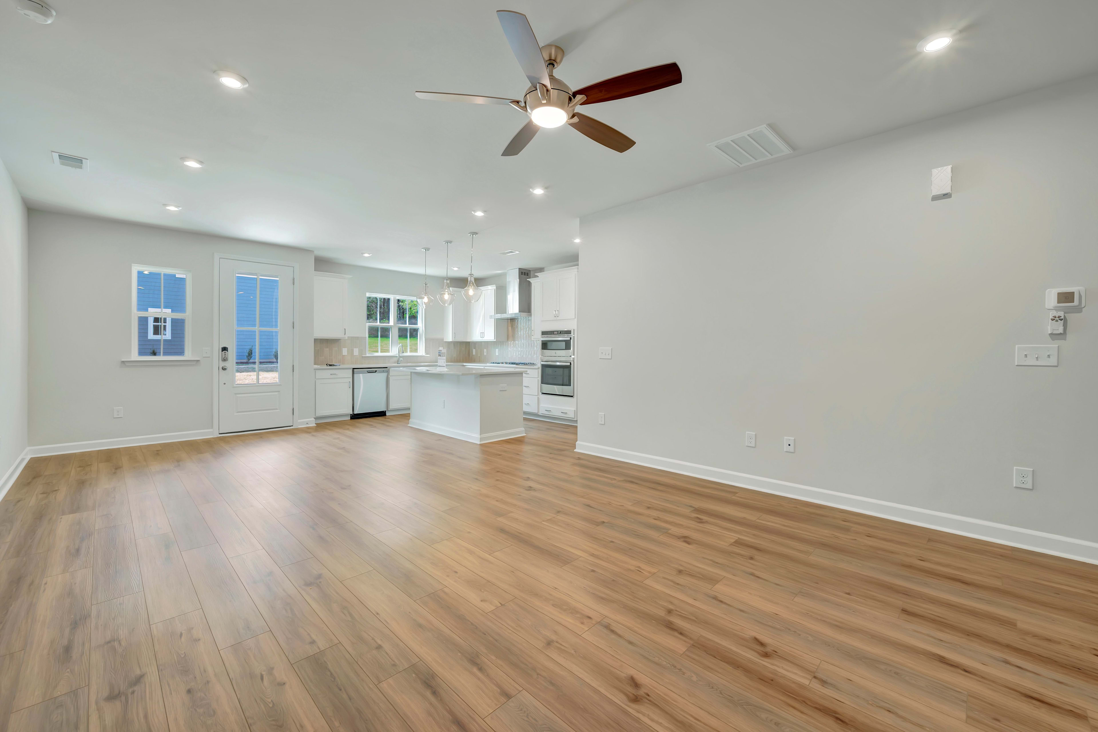 A spacious, open-concept kitchen and living room with white walls, hardwood floors, and a ceiling fan.
