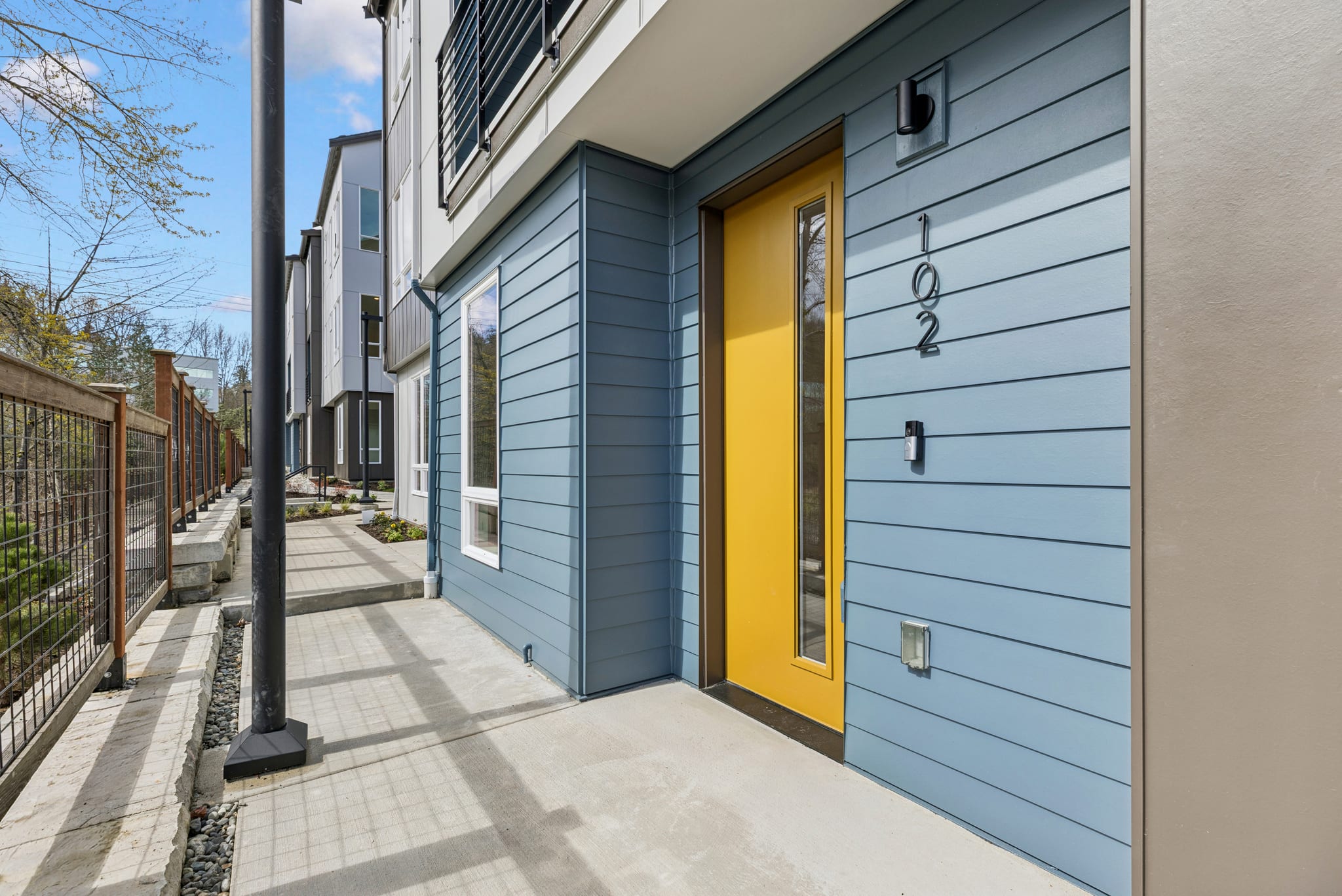 A row of modern townhouses with blue siding, yellow front doors, and a paved walkway leading past them.