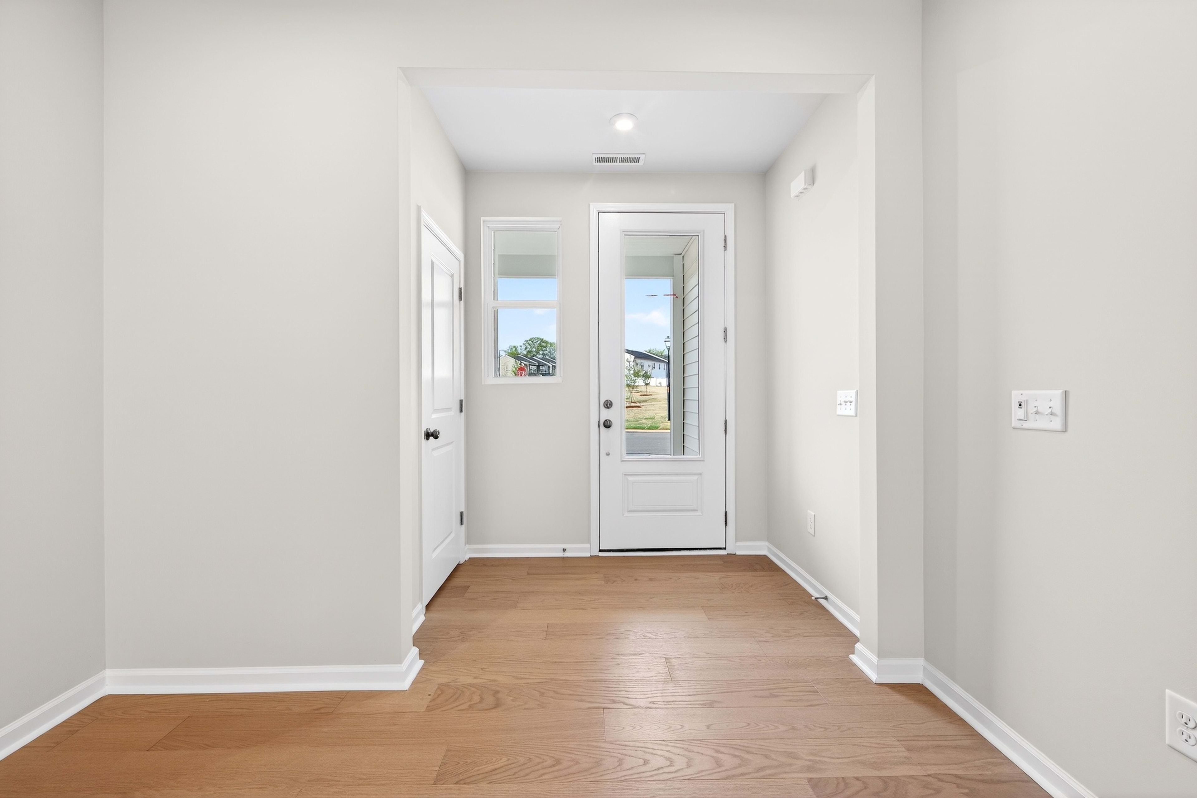 A bright and airy hallway with a white door leading to the outdoors, featuring a wooden floor and neutral-colored walls.