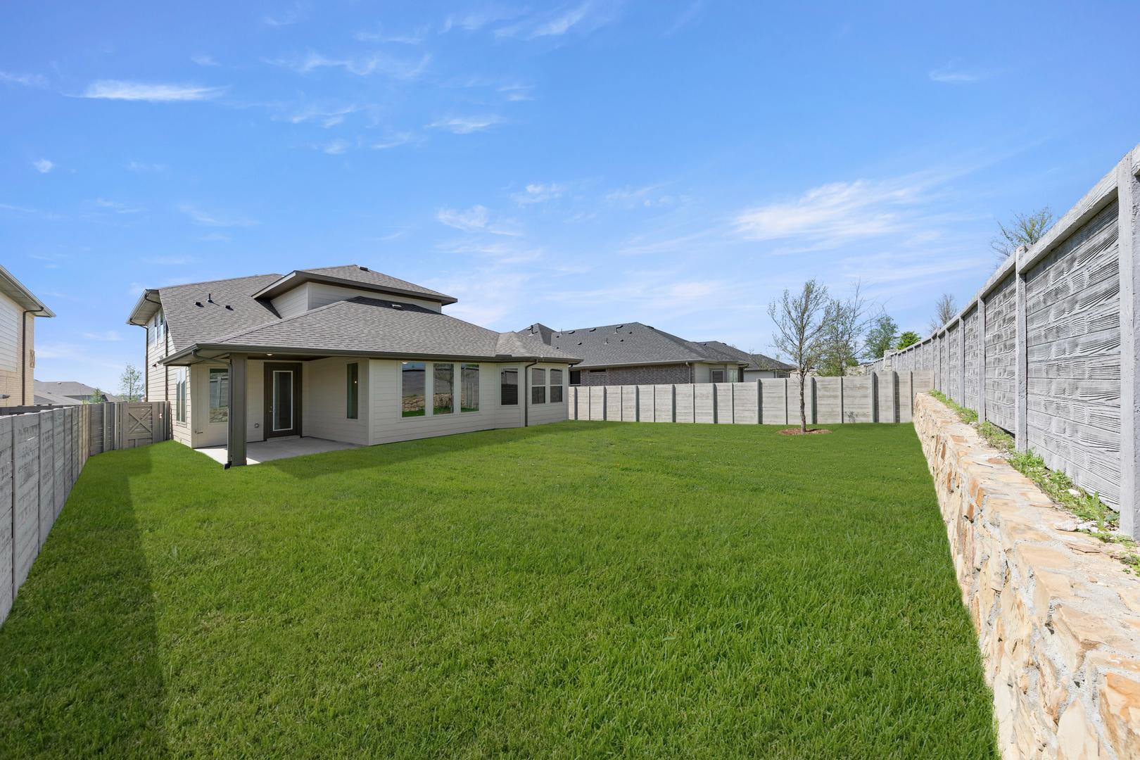A well-manicured lawn surrounds a modern, two-story house with a stone exterior, set against a clear blue sky with scattered clouds.