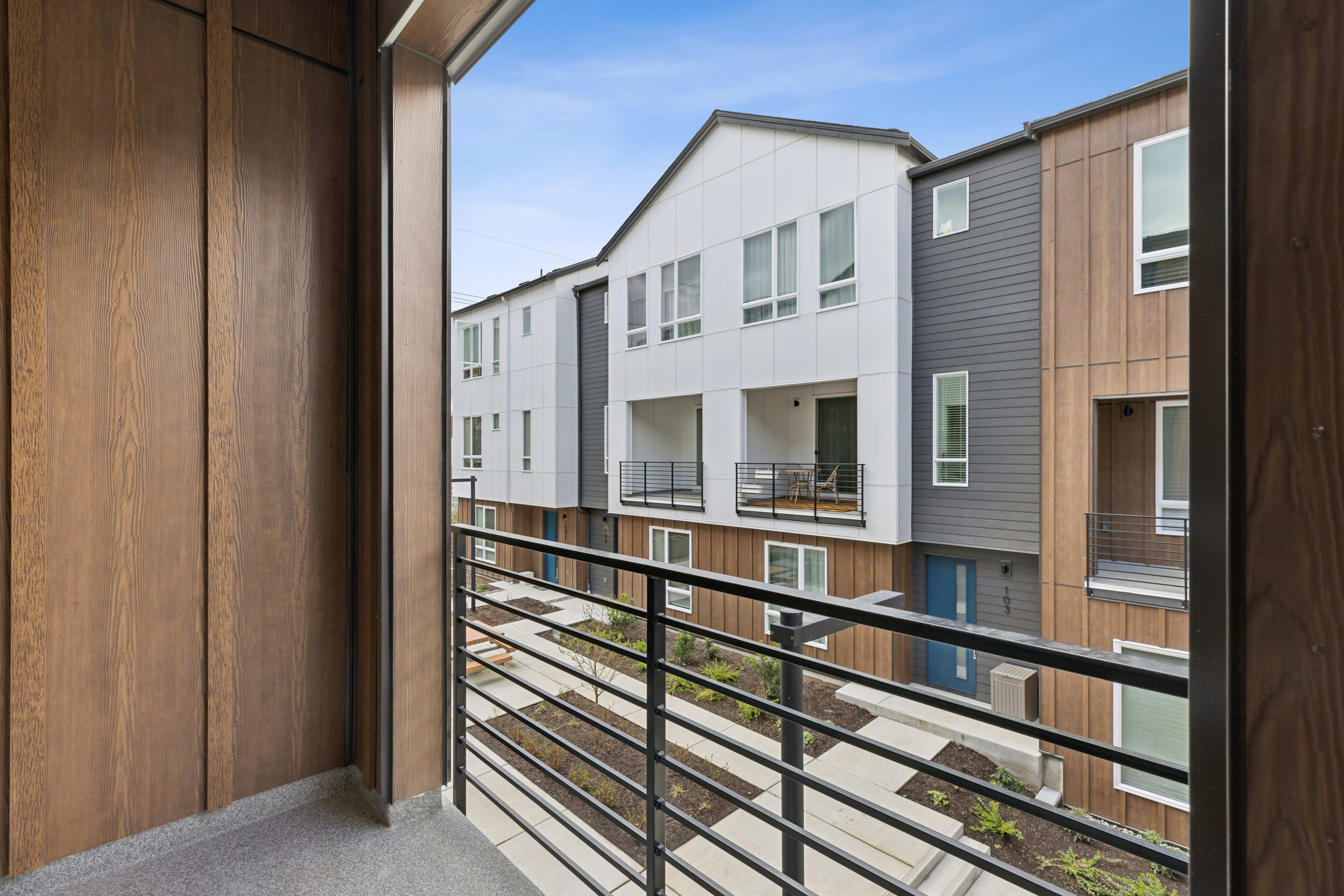 A wooden balcony with a metal railing overlooks a row of modern, multi-story residential buildings with a mix of siding materials and window styles.