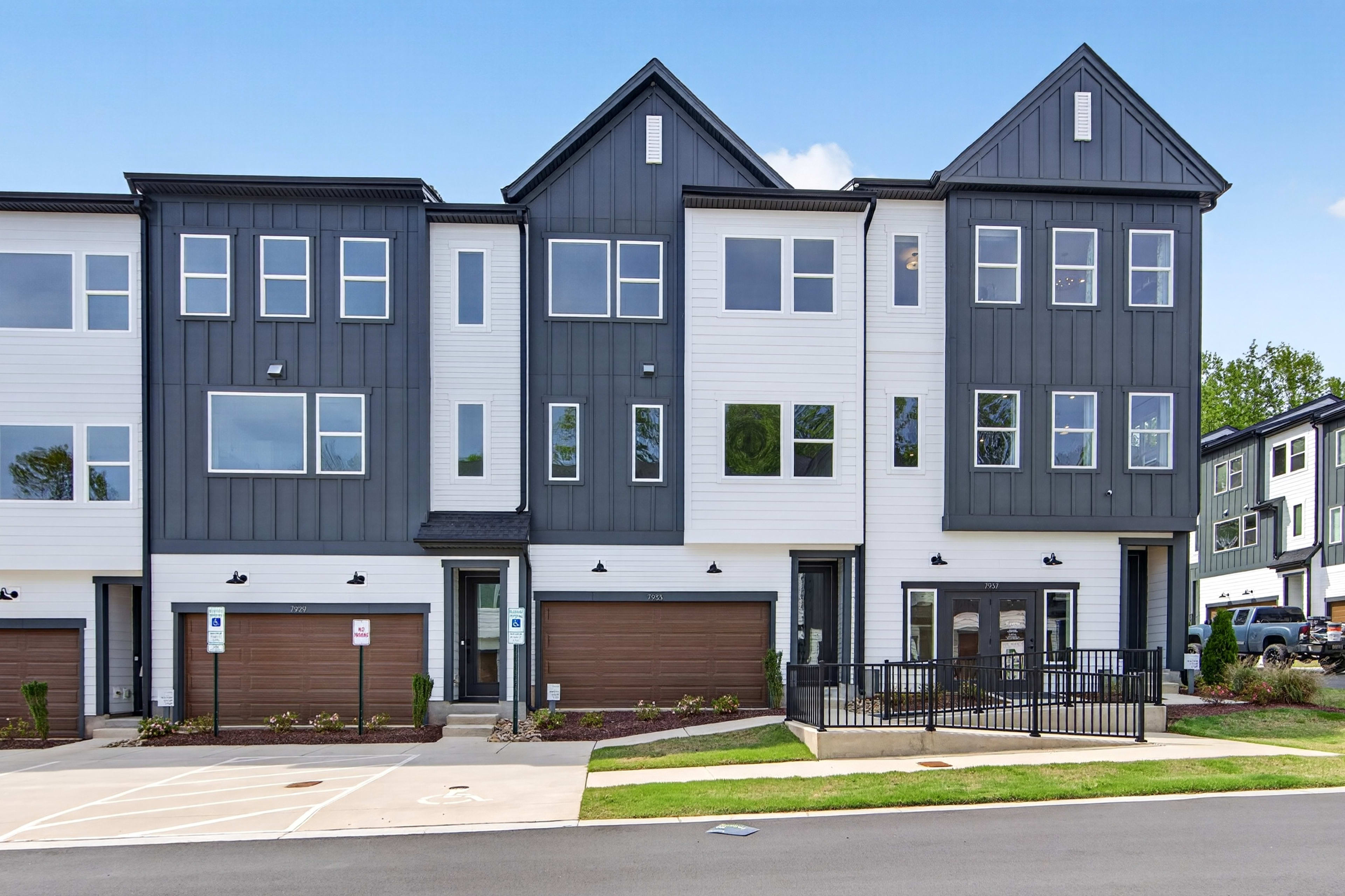A modern, multi-story townhouse complex with gray siding, gabled roofs, and attached garages, set against a backdrop of a clear blue sky and lush greenery.