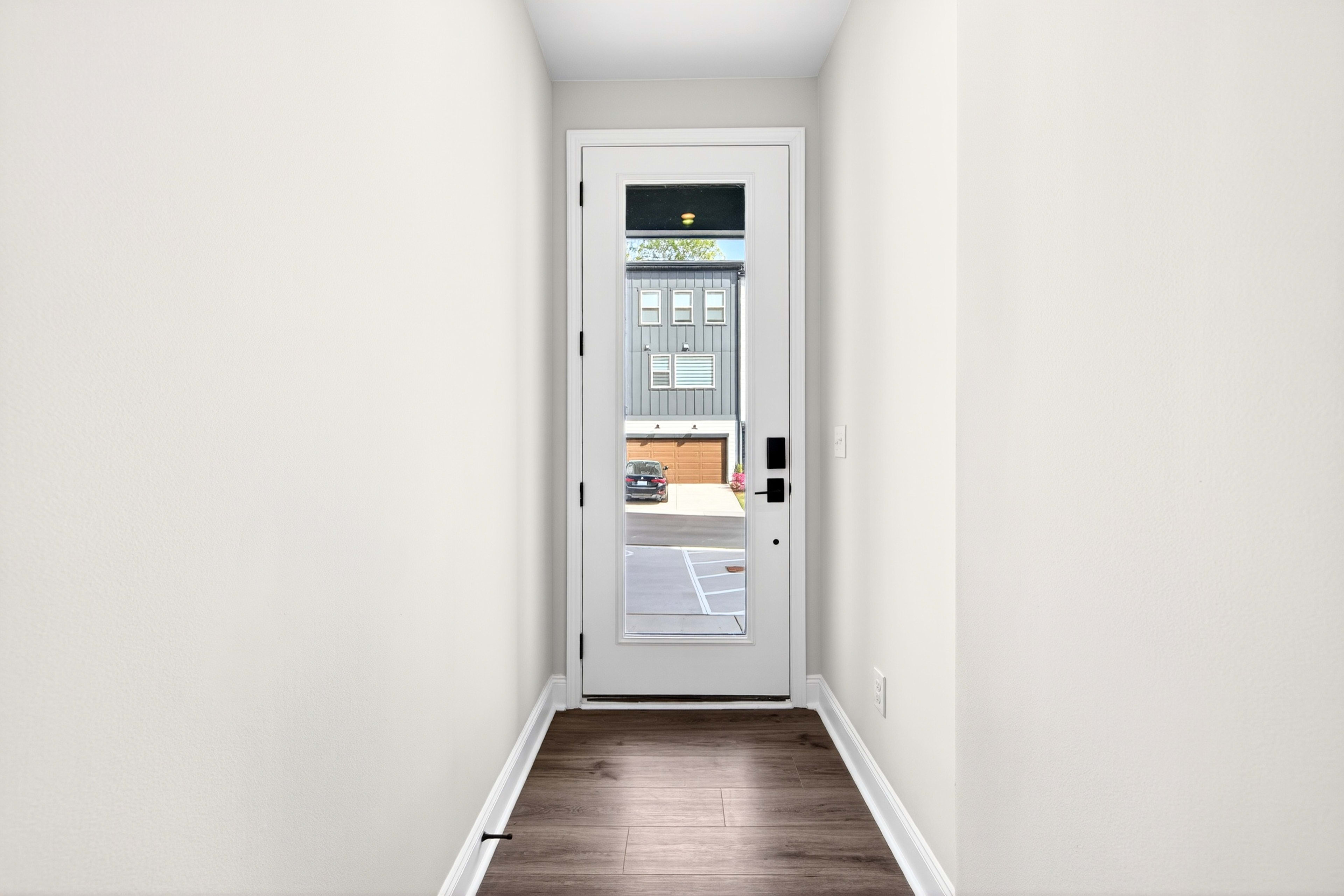 A narrow white hallway with a wooden floor and a white door at the end, partially open and revealing a room beyond.