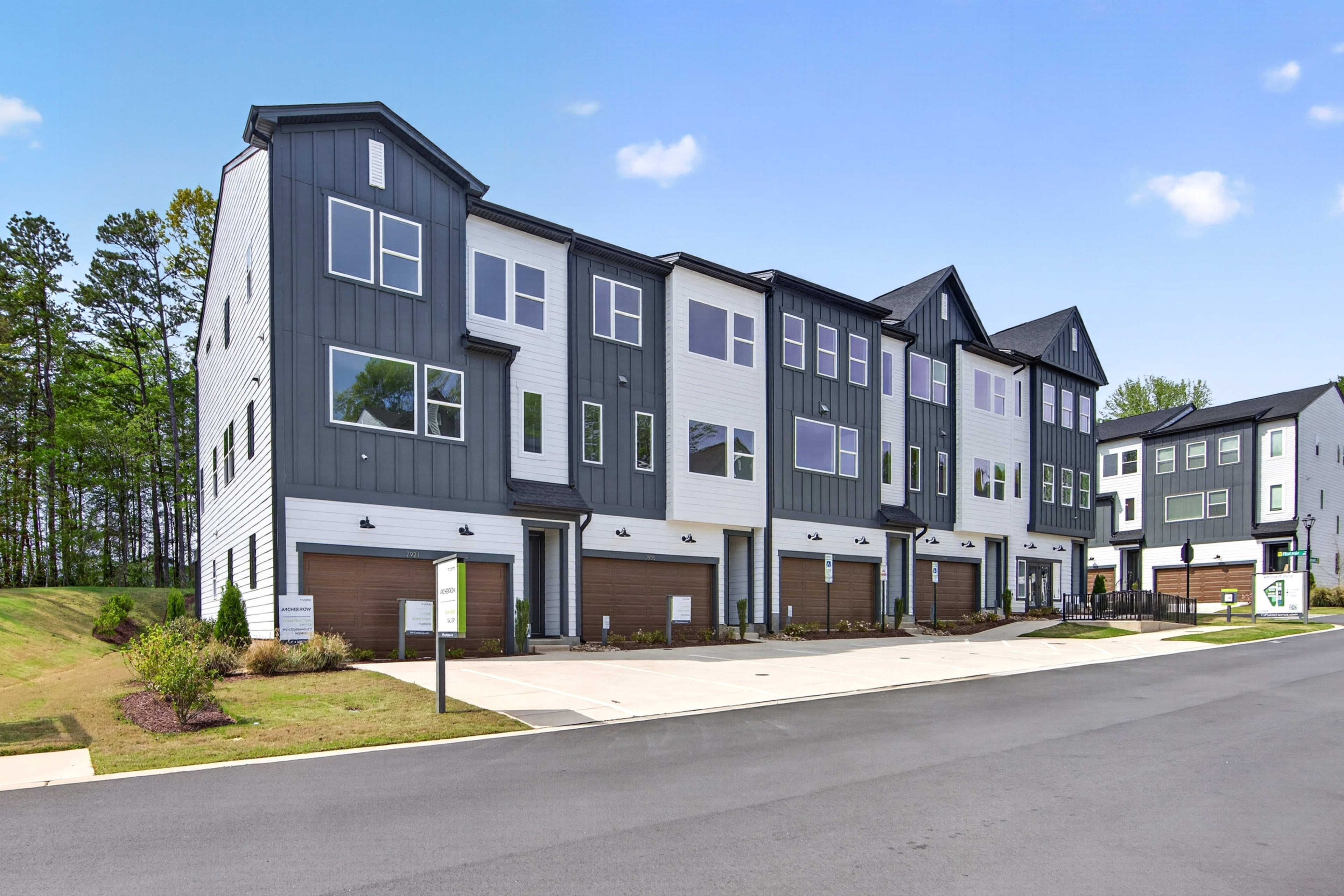 A row of modern, multi-story townhouses with dark gray siding and large windows, surrounded by trees and a paved road in the foreground.