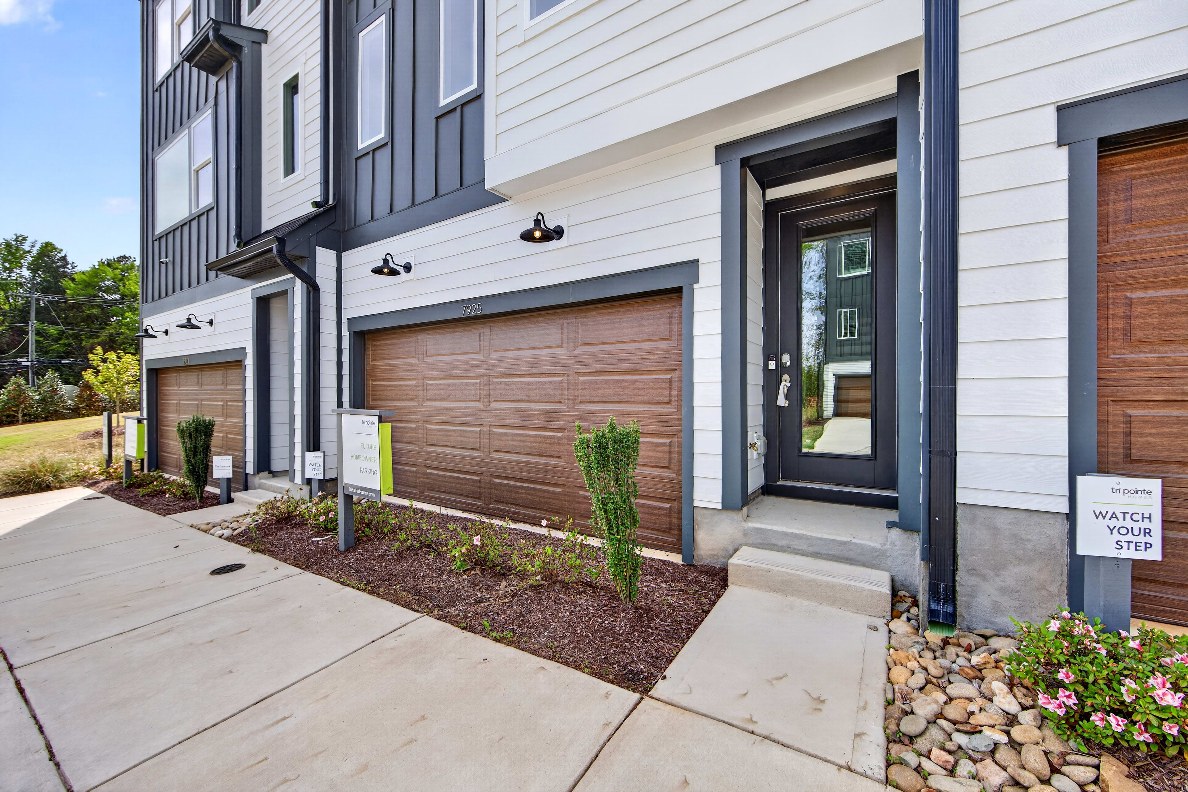 The image depicts a modern, multi-story residential building with a garage door, landscaping, and a walkway leading to the entrance.