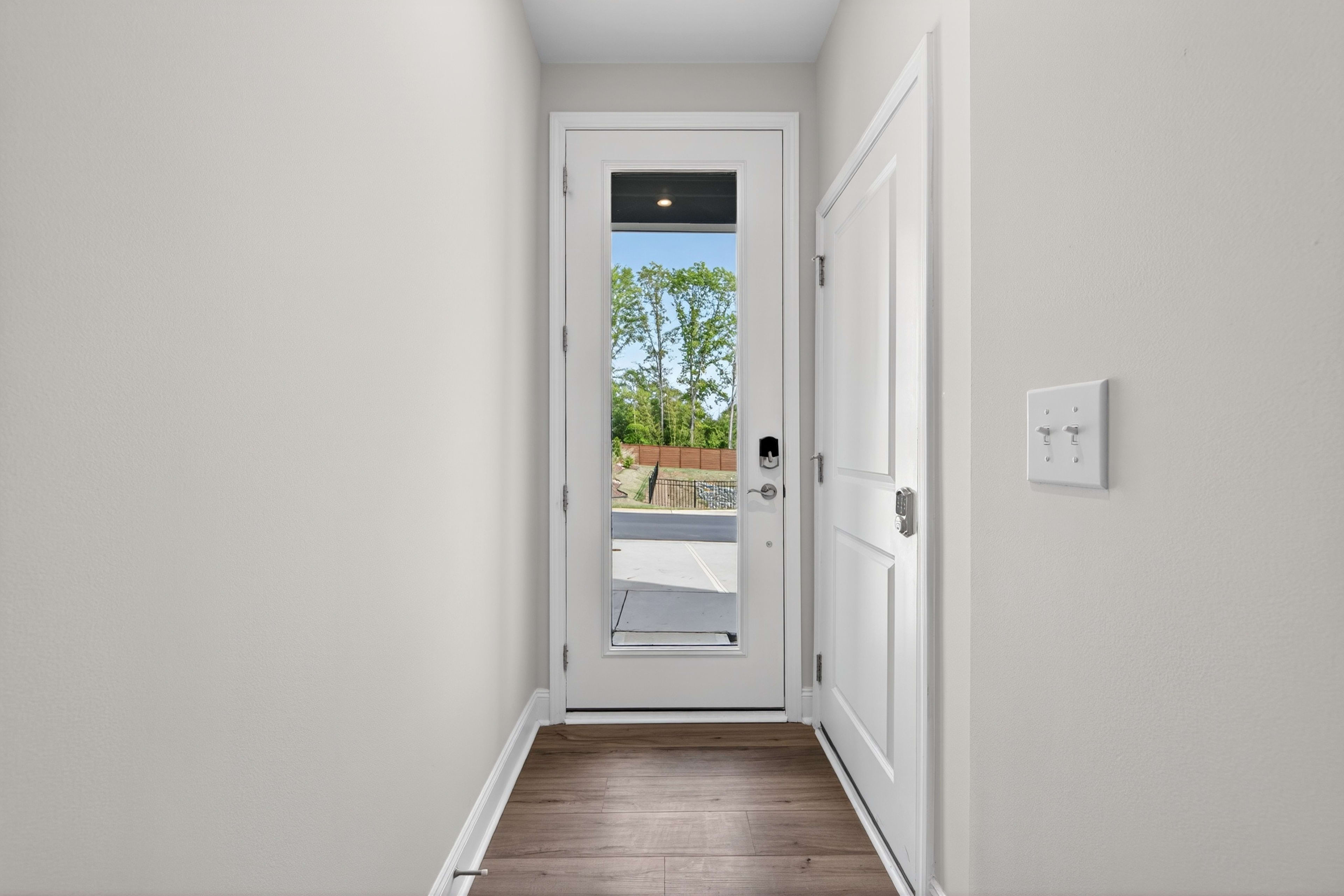 A bright, airy hallway with a white door leading to a view of lush greenery outside.