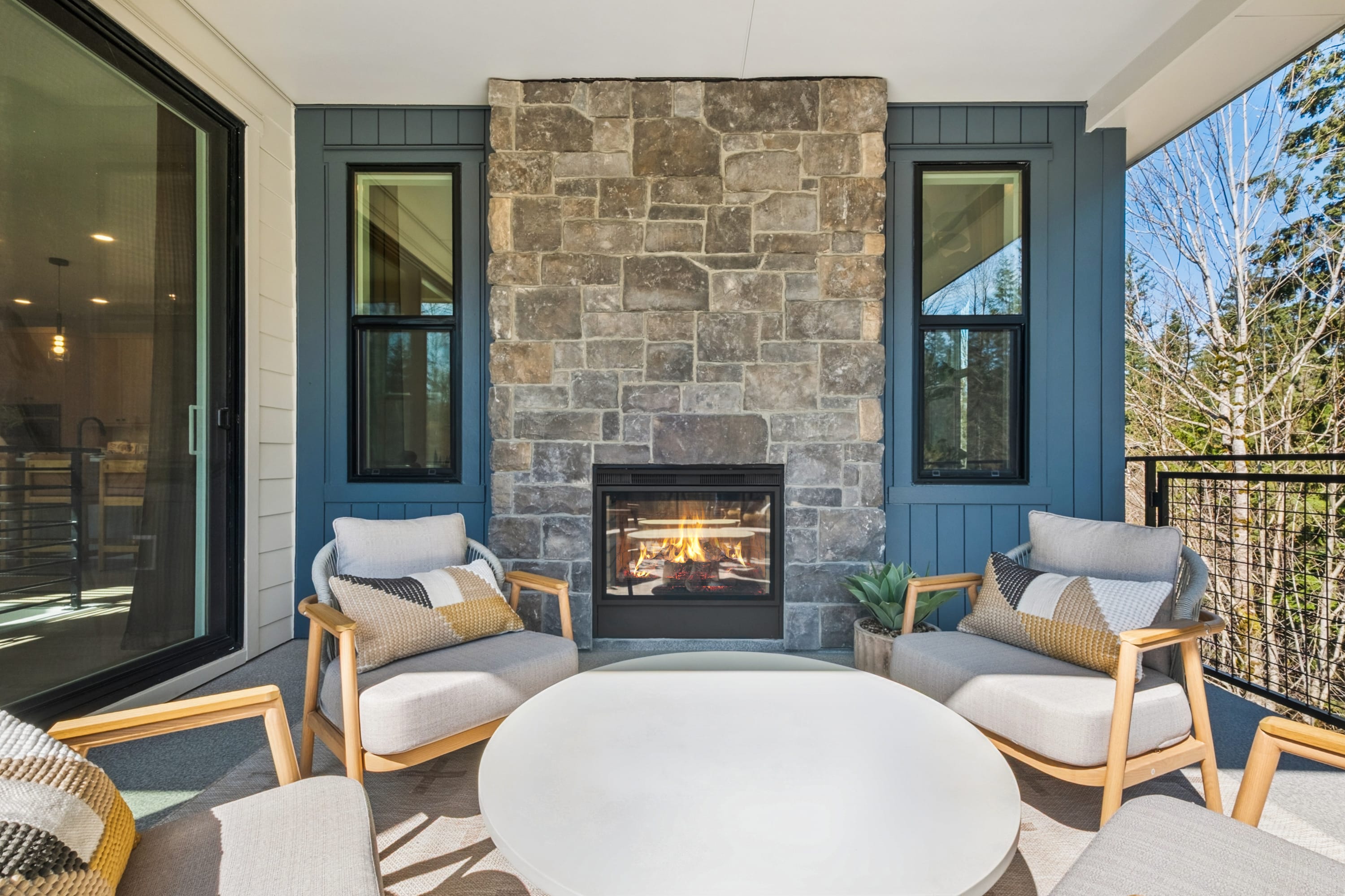 A cozy outdoor seating area with a stone fireplace, comfortable chairs, and a round table, surrounded by large windows and a scenic natural backdrop.