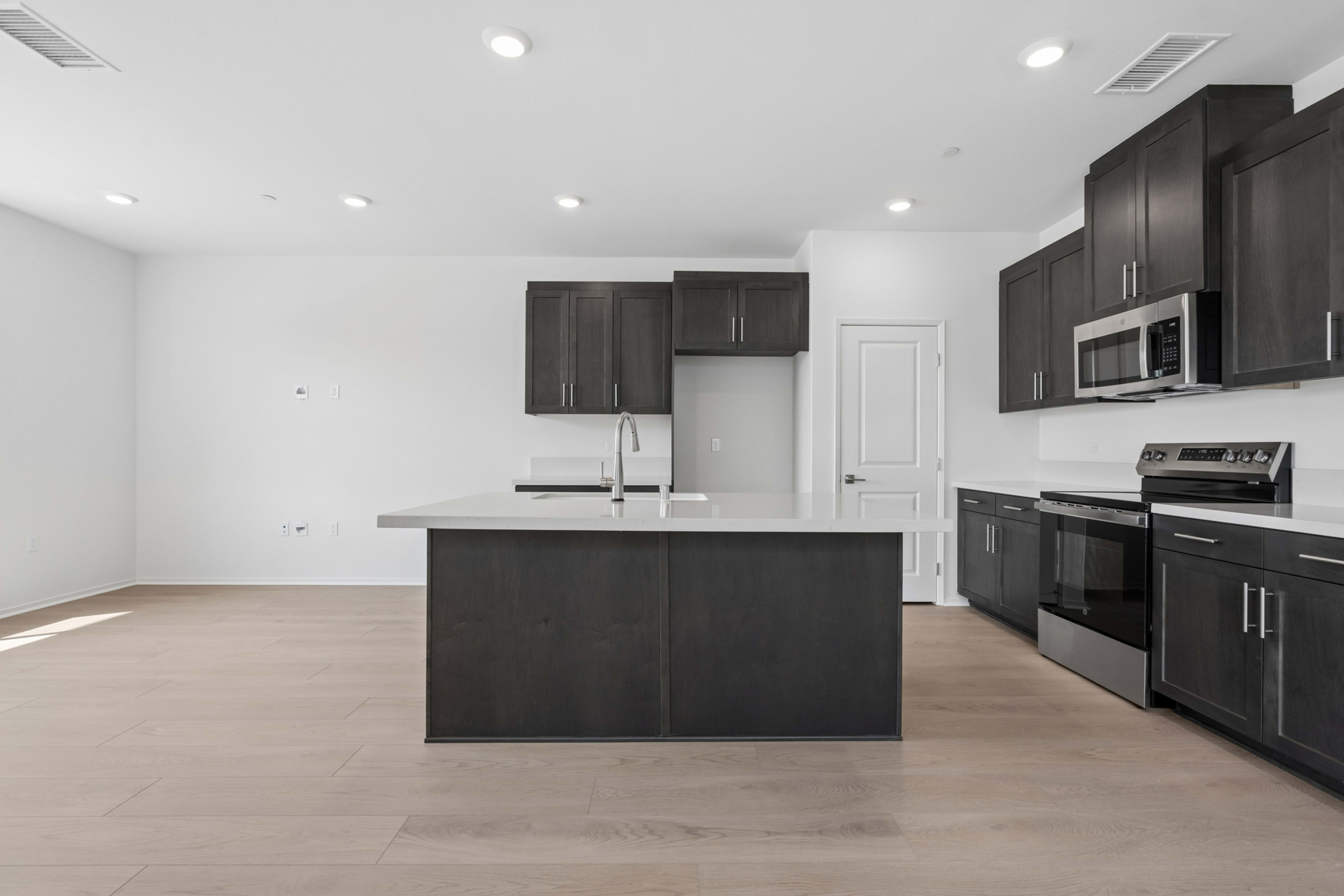 A modern, open-concept kitchen with dark cabinets, a central island, and stainless steel appliances, set against a bright, minimalist background.