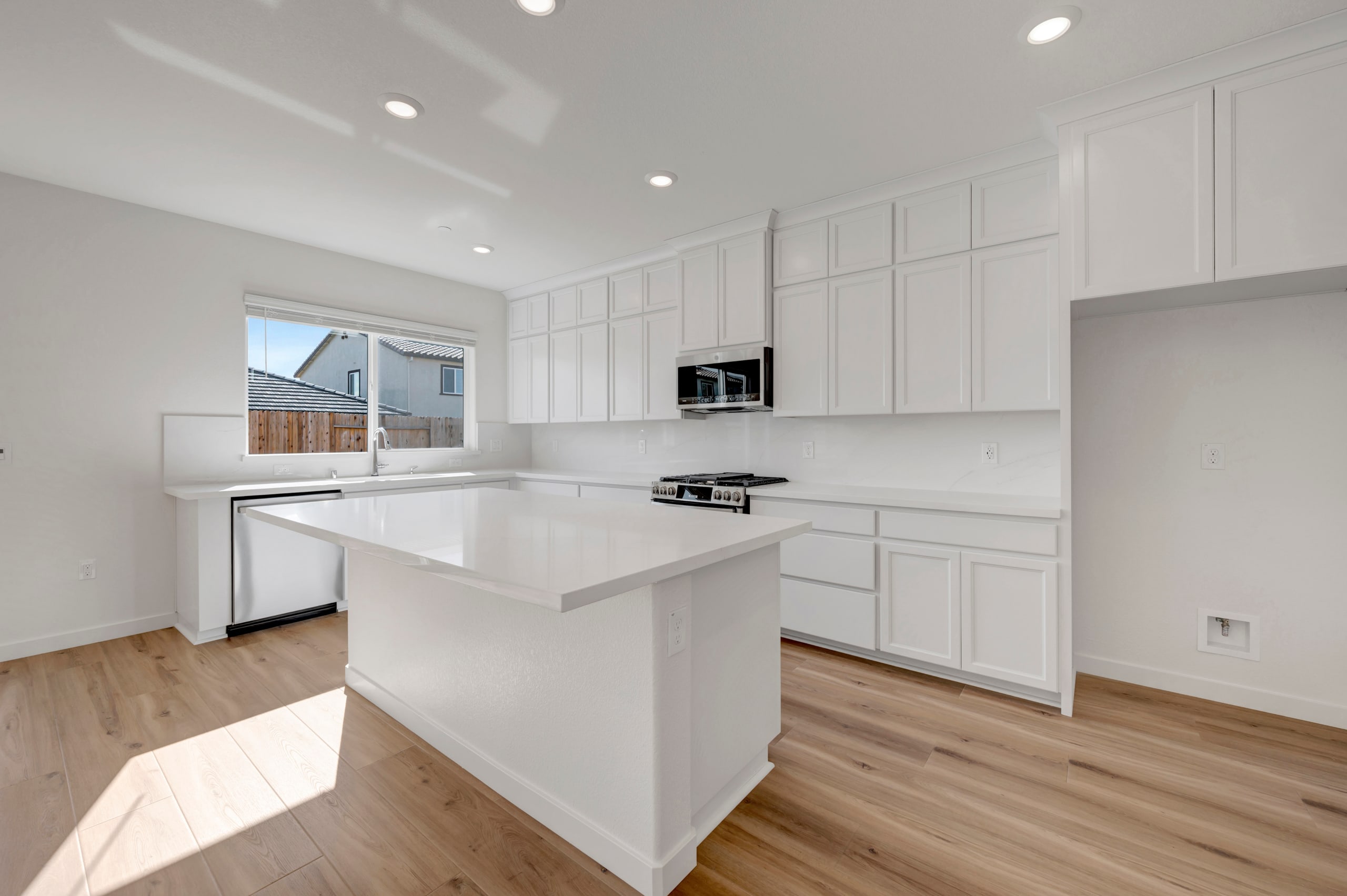 A modern, bright kitchen with white cabinets, a white countertop, and hardwood floors, with a window visible in the background.