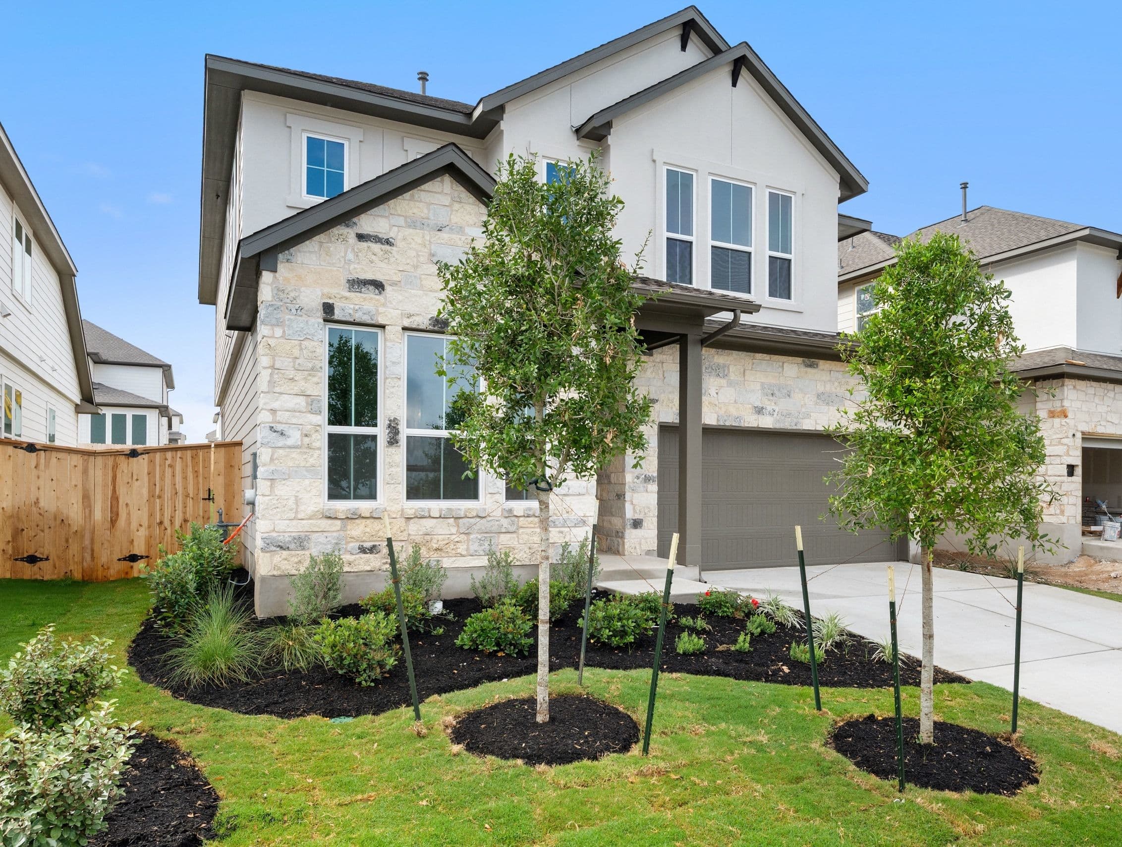 A two-story residential house with a stone exterior, surrounded by a well-manicured lawn and landscaping, including young trees and shrubs, set against a clear blue sky.