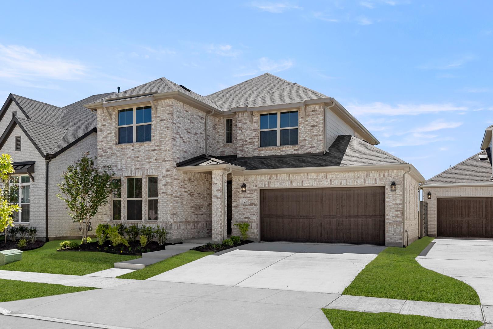 A two-story residential house with a stone exterior, a garage, and a well-manicured lawn in the foreground, set against a clear blue sky with some clouds.