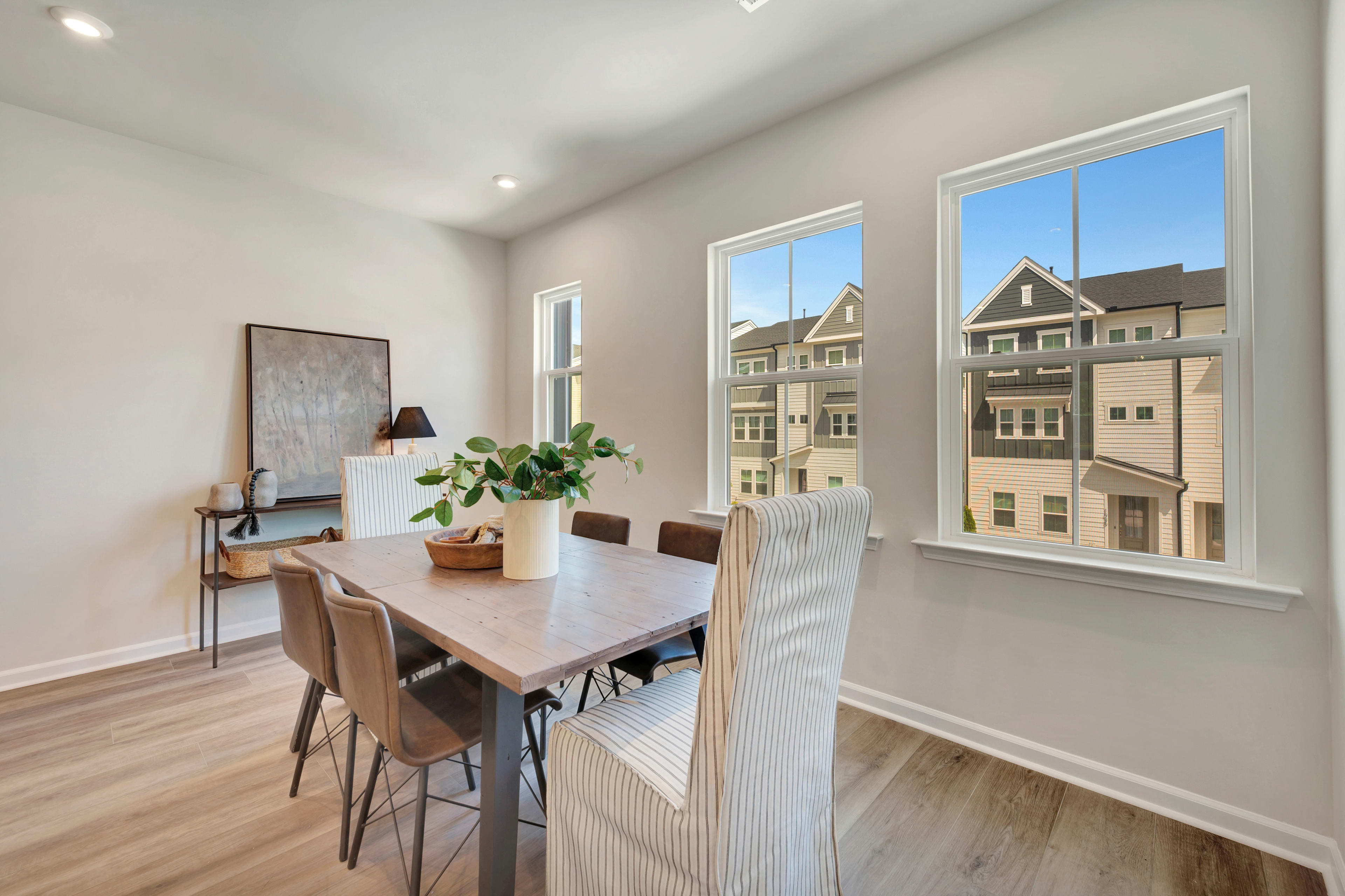 A modern and bright dining room with a wooden table, chairs, and a large window overlooking a residential neighborhood.