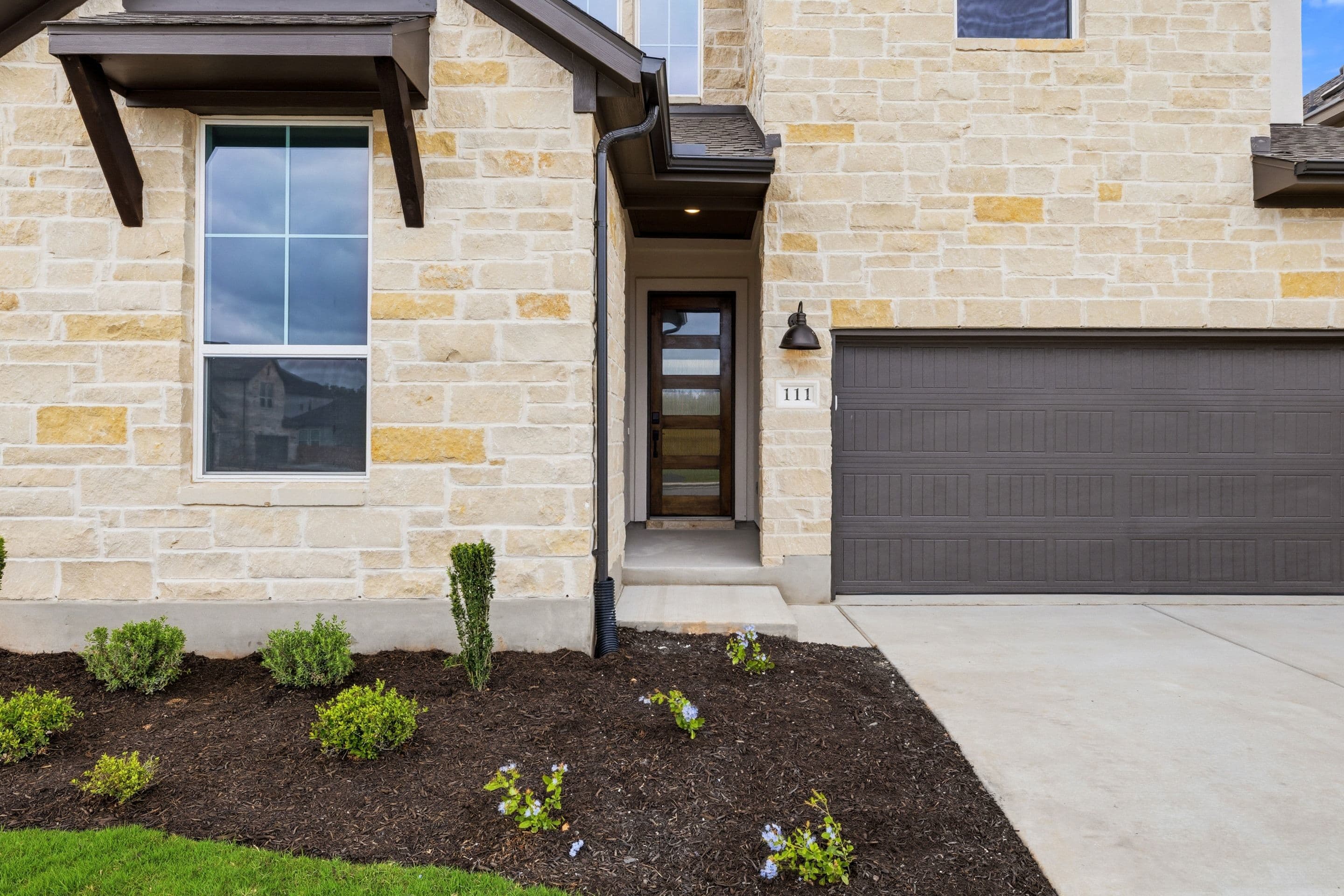 A modern, stone-clad house with a garage door, surrounded by a freshly landscaped yard with small plants and shrubs.
