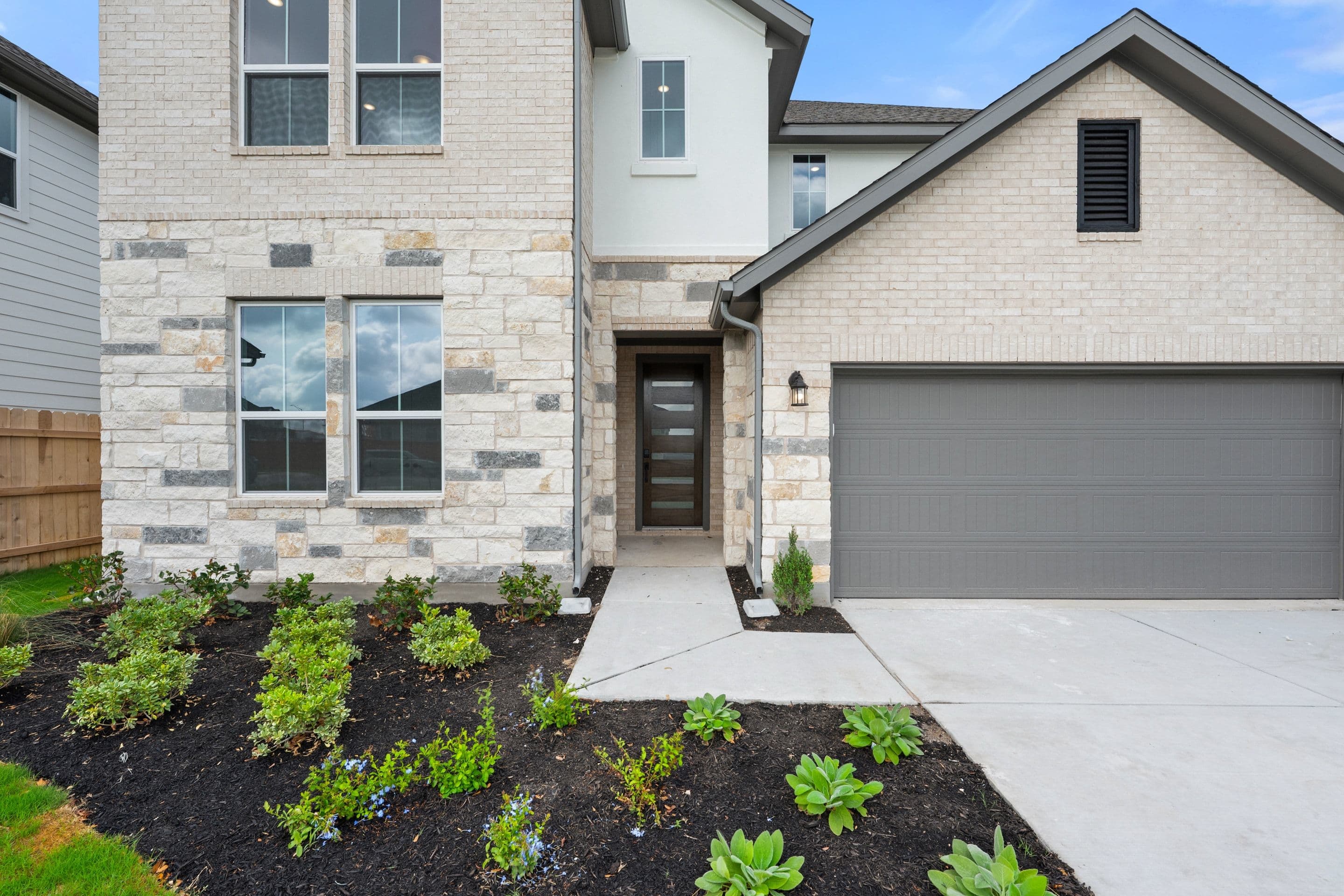 A modern two-story house with a stone and siding exterior, a gray garage door, and a well-landscaped front yard with lush greenery.