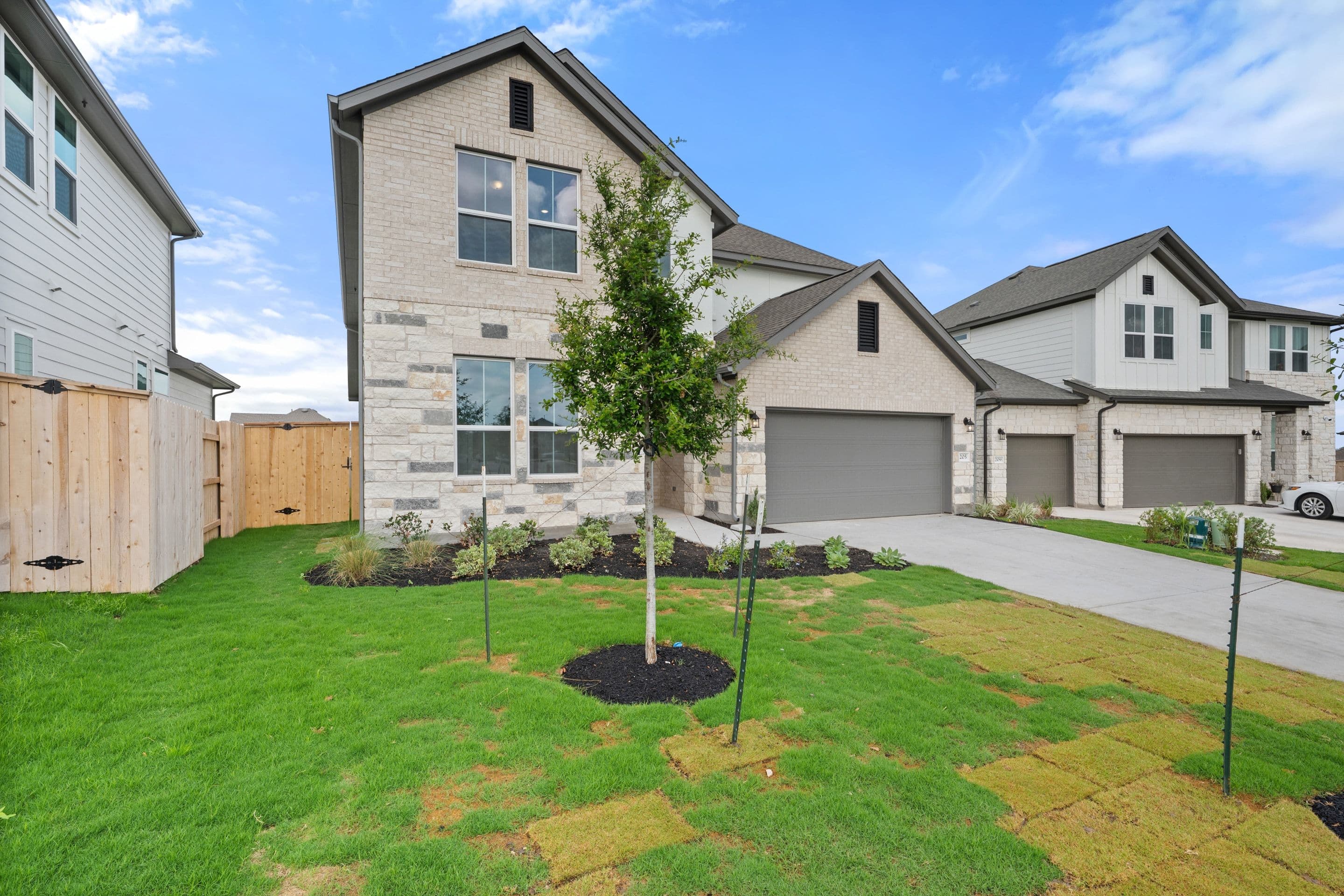 A newly constructed residential neighborhood with modern, two-story homes featuring gray siding, large windows, and attached garages. The foreground showcases a well-manicured lawn with a newly planted tree.