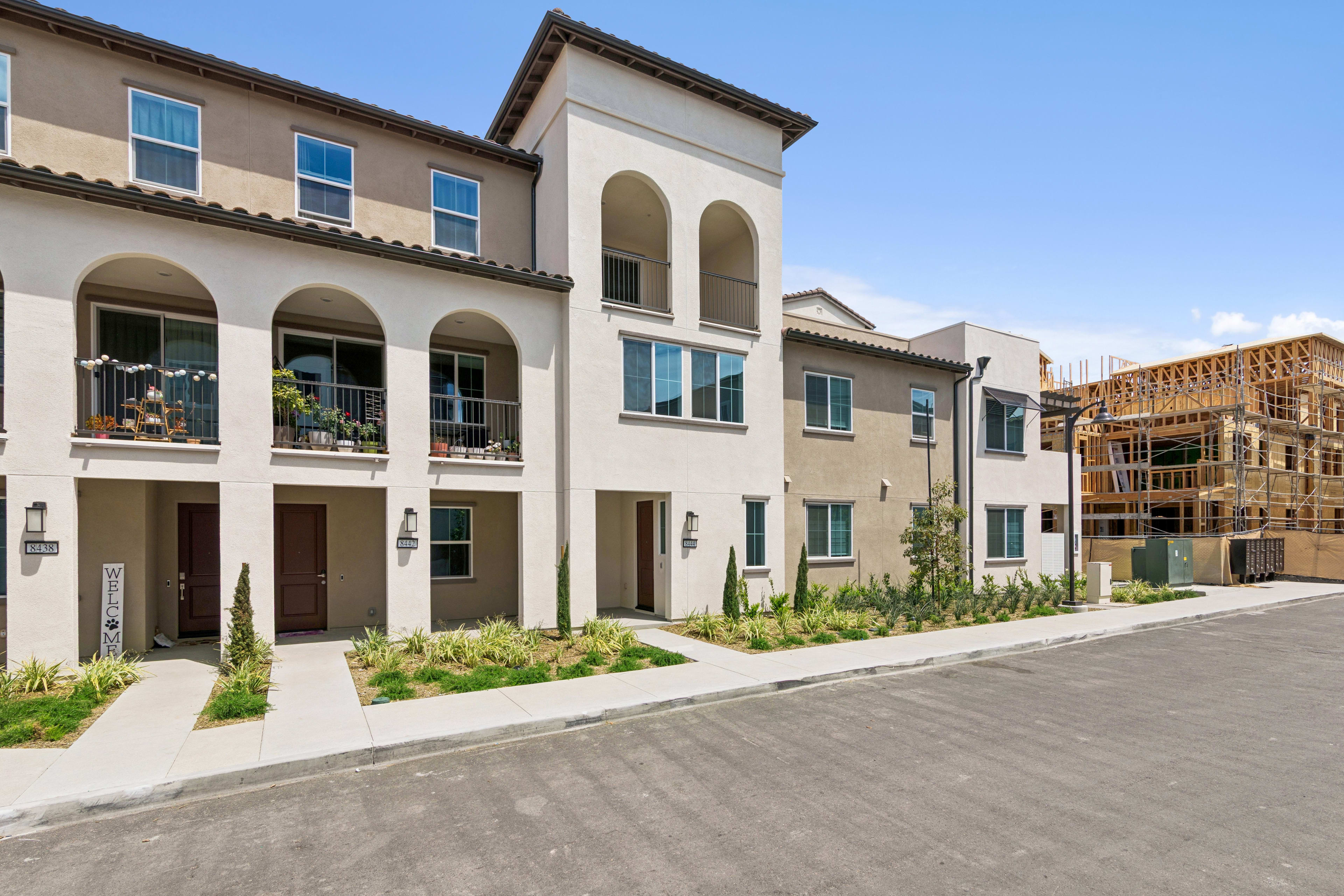 The image depicts a row of multi-story residential buildings with a Mediterranean-style architectural design, featuring arched entryways, balconies, and a well-maintained landscaped area in the foreground.
