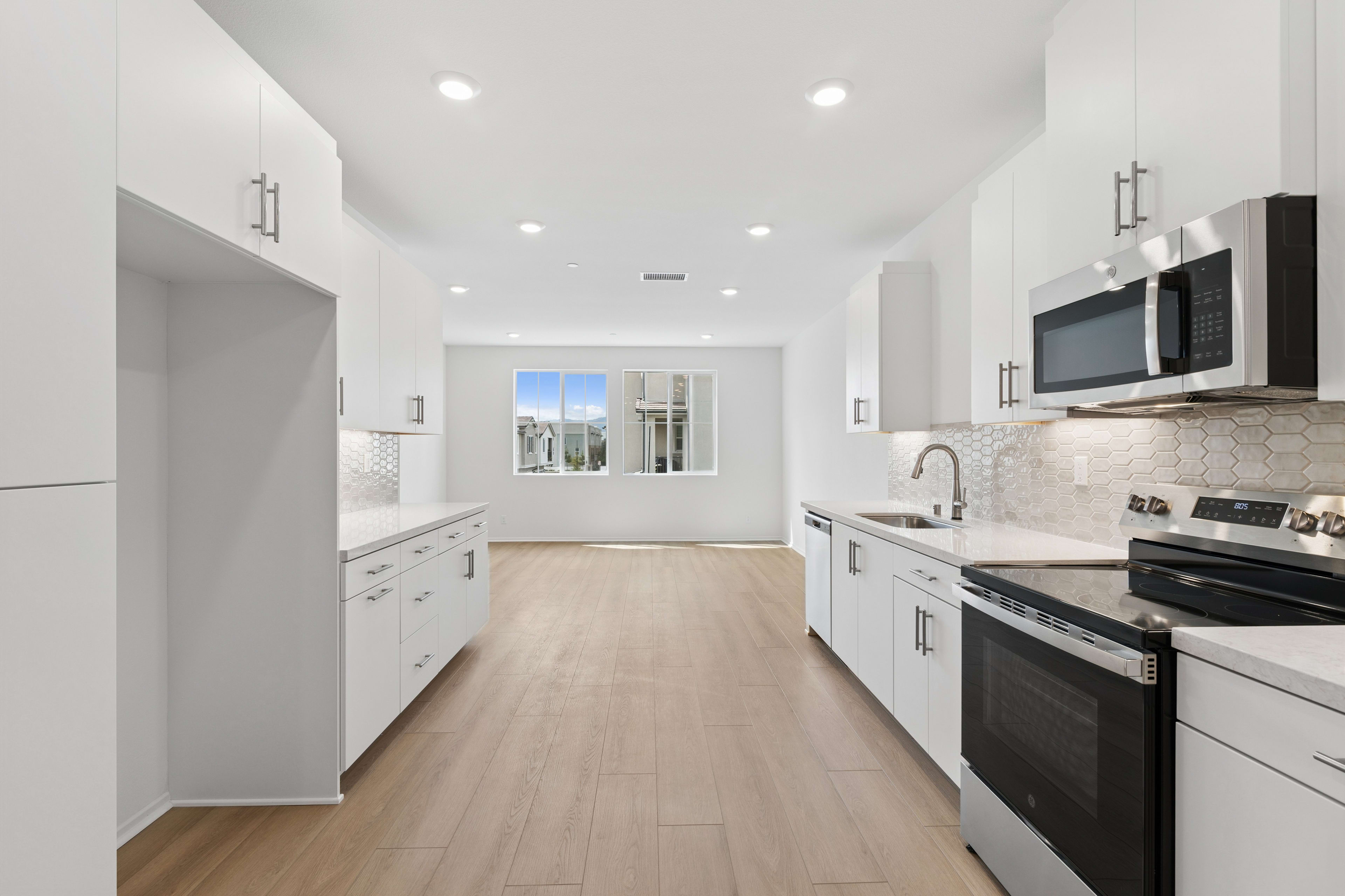A modern, bright, and spacious kitchen with white cabinets, stainless steel appliances, and a hardwood floor leading to a window with a view of the outdoors.