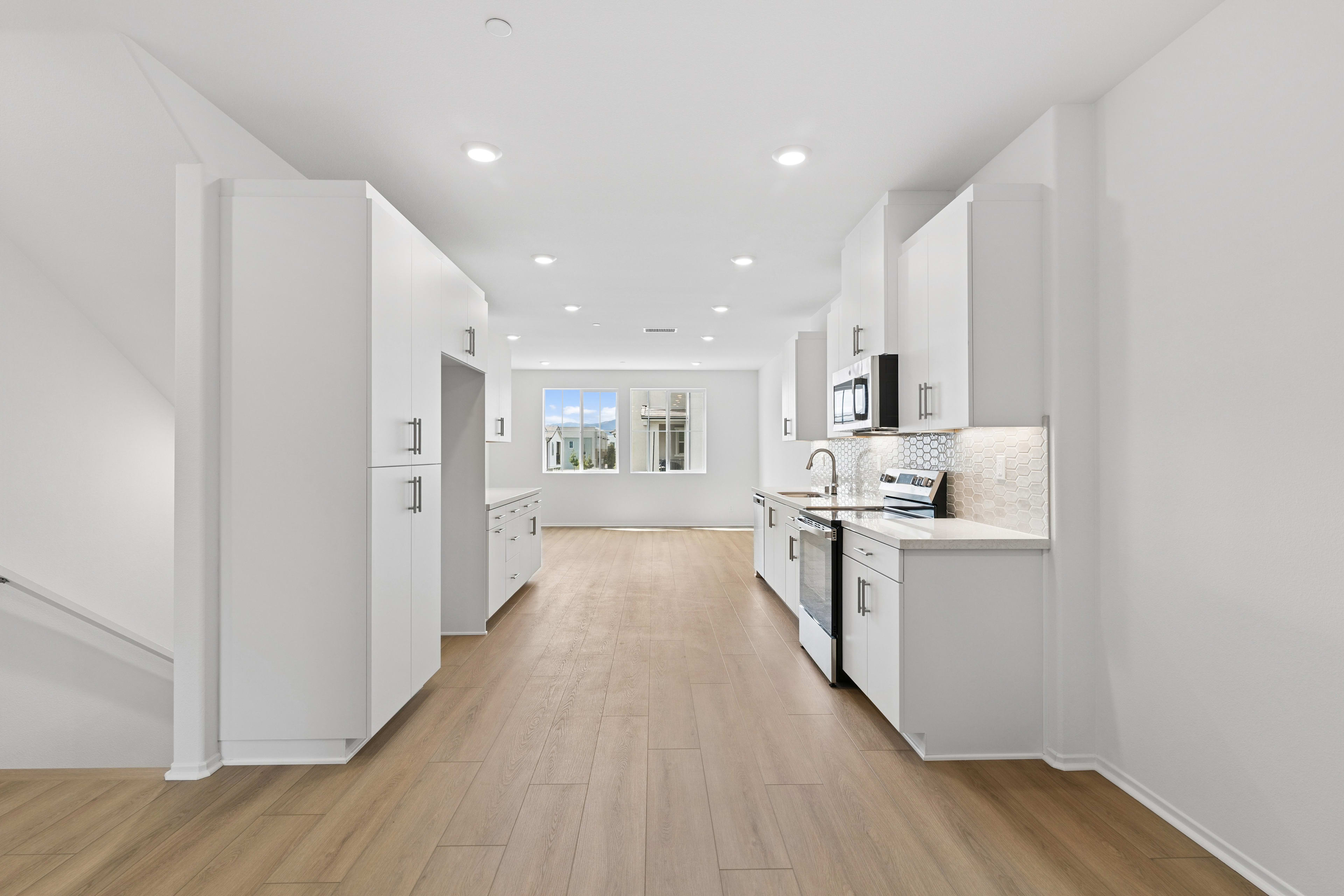 A bright, spacious hallway with a kitchen visible at the end, featuring white walls, hardwood floors, and recessed lighting.